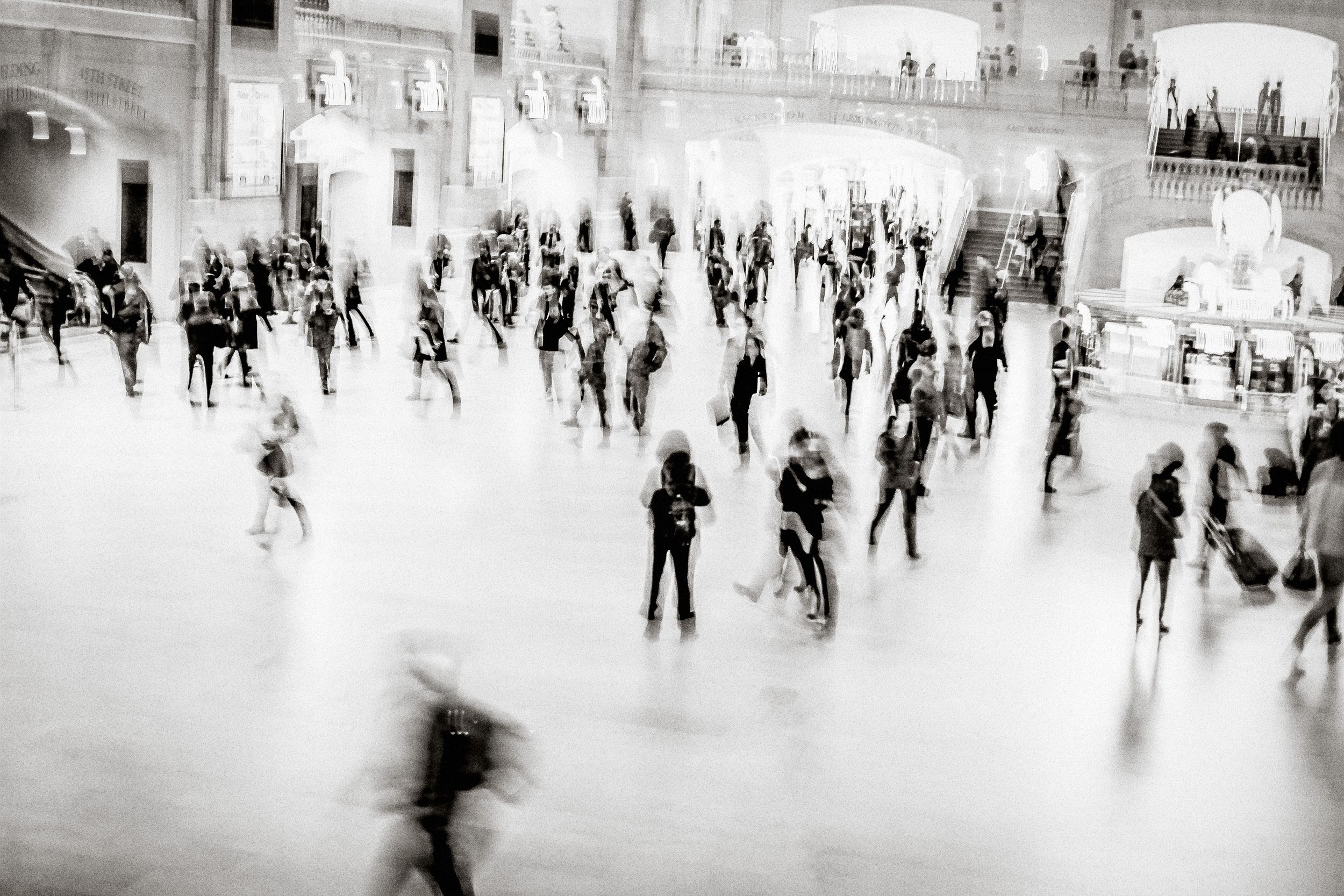 Blurred black and white photo of crowds of people moving through a grand busy public hall or train station.