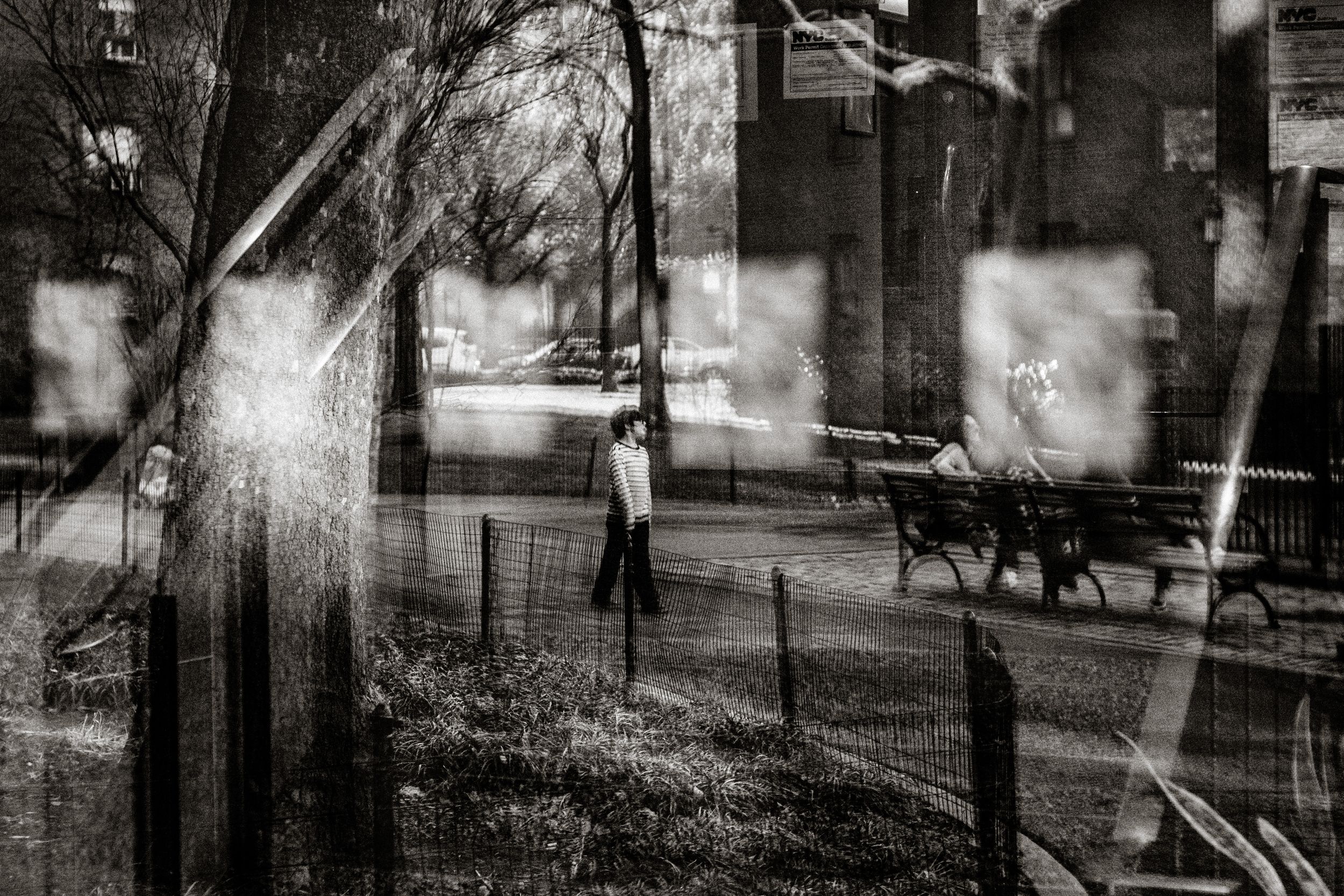 Black and white double exposure street photo showing a lone figure walking near a park fence with benches and bare trees.