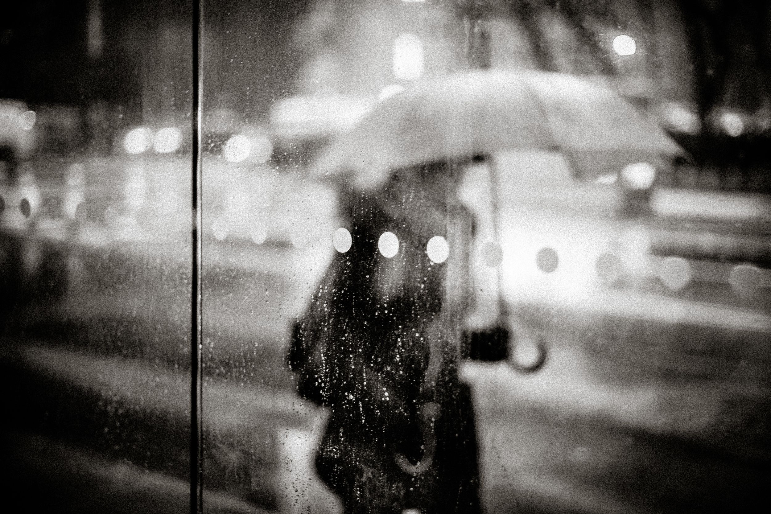 Black and white photo of a person holding umbrella in rain at night, city lights bokeh in background.