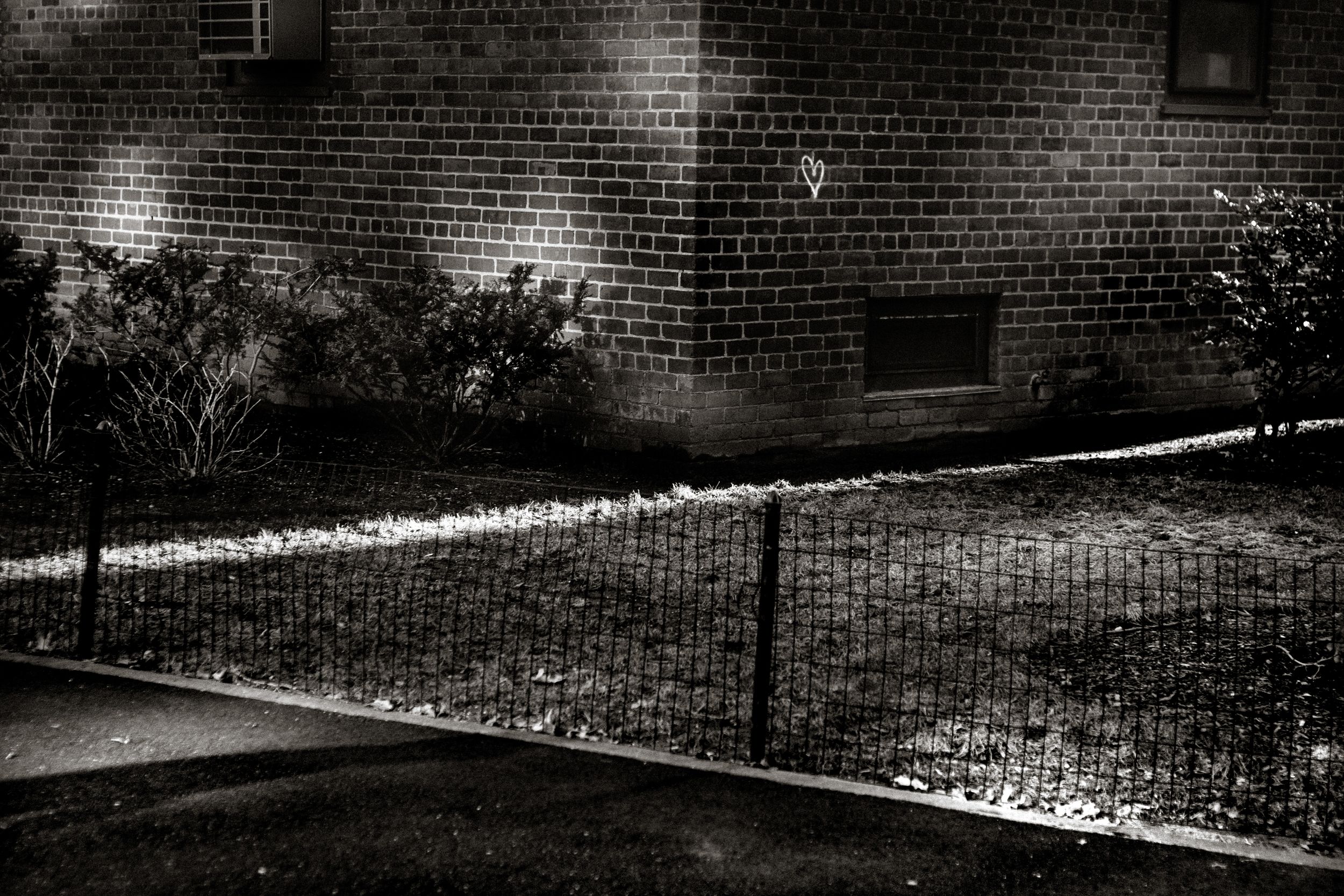 Black and white night photo of brick building with chain-link fence, illuminated shrubs, and cobblestone ground.
