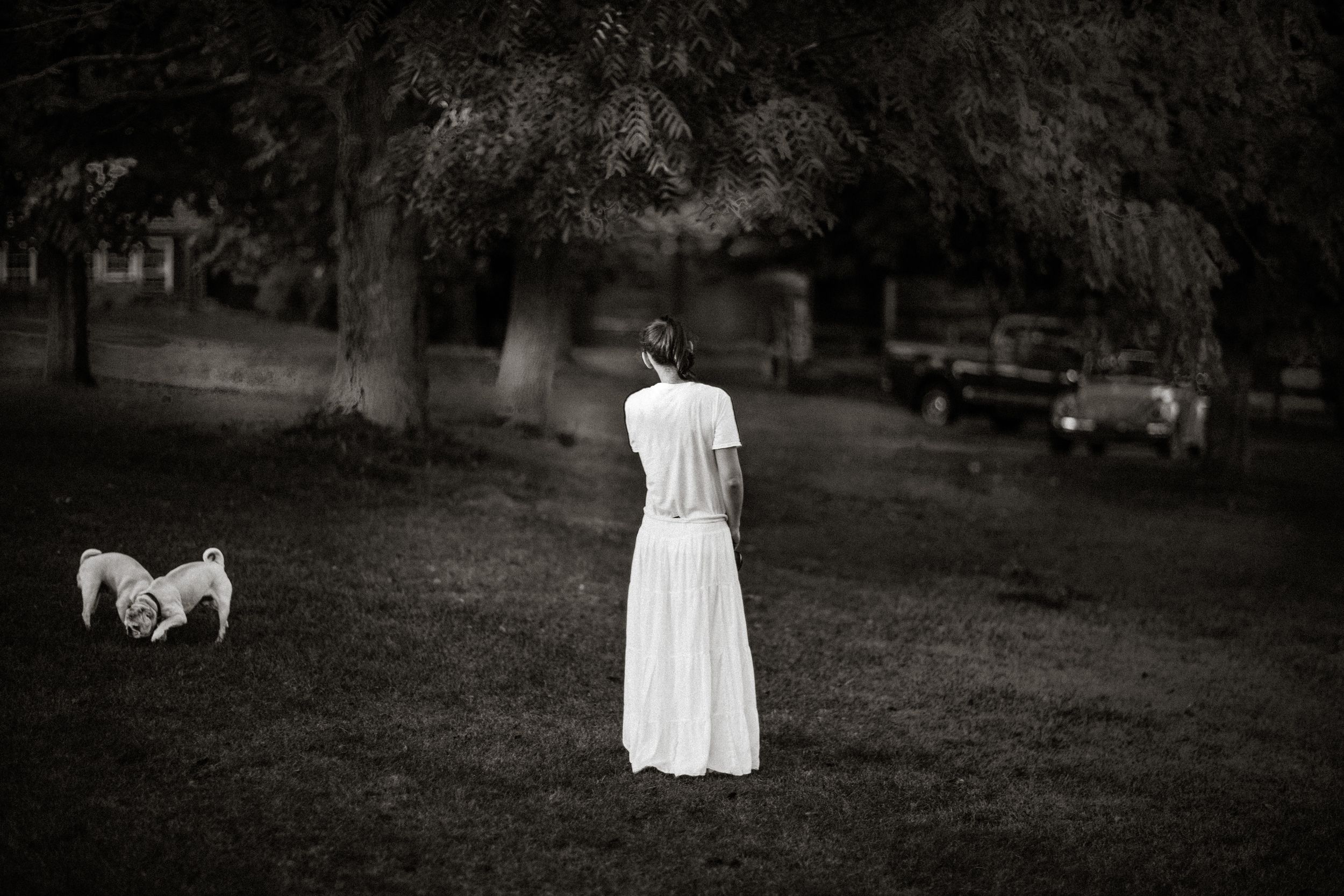 Woman in white dress stands in park facing small dog near vintage car in black and white photo.