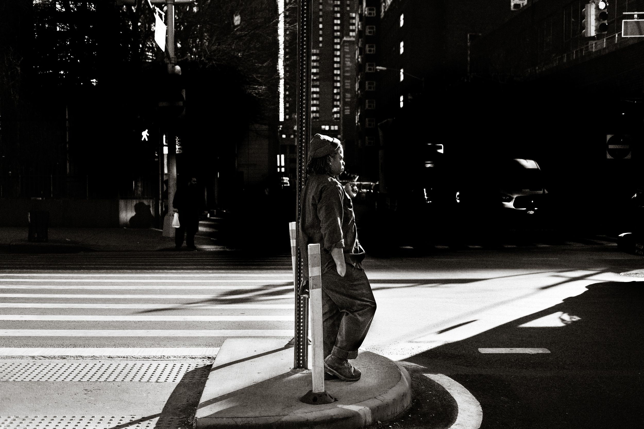 Black and white photo of a person standing by a pole at a city crosswalk, with dramatic light and shadow.