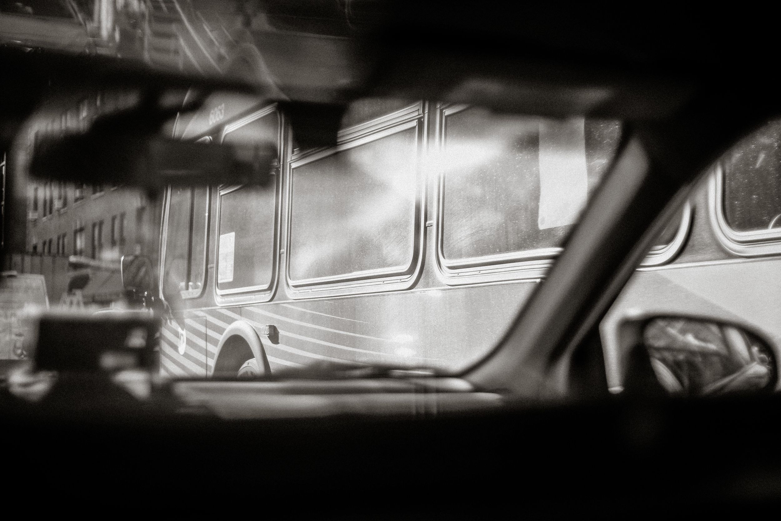 Black and white photo of a bus viewed through a car window, capturing an urban street scene in moody monochrome.