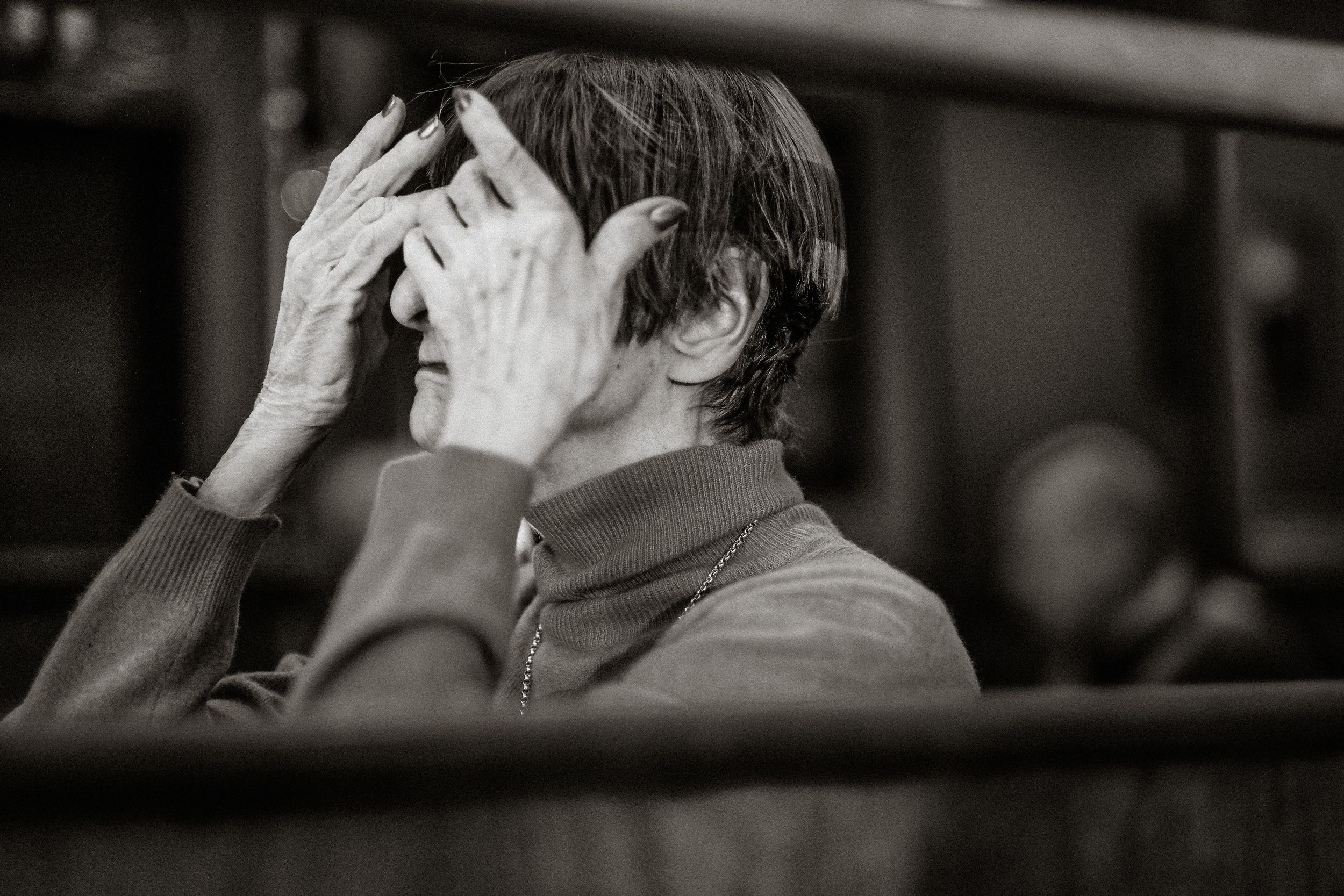 Black and white photo of a stressed man covering his face with both hands while sitting in a wooden pew.