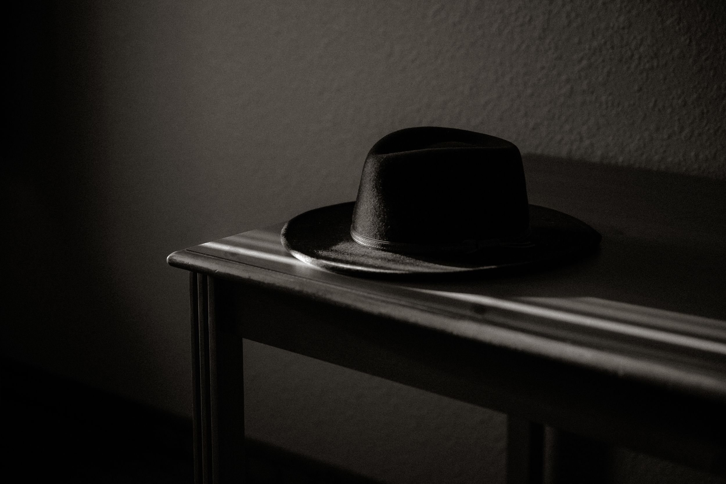 A dark black fedora hat resting on a wooden table in a dimly lit room with dramatic shadows.