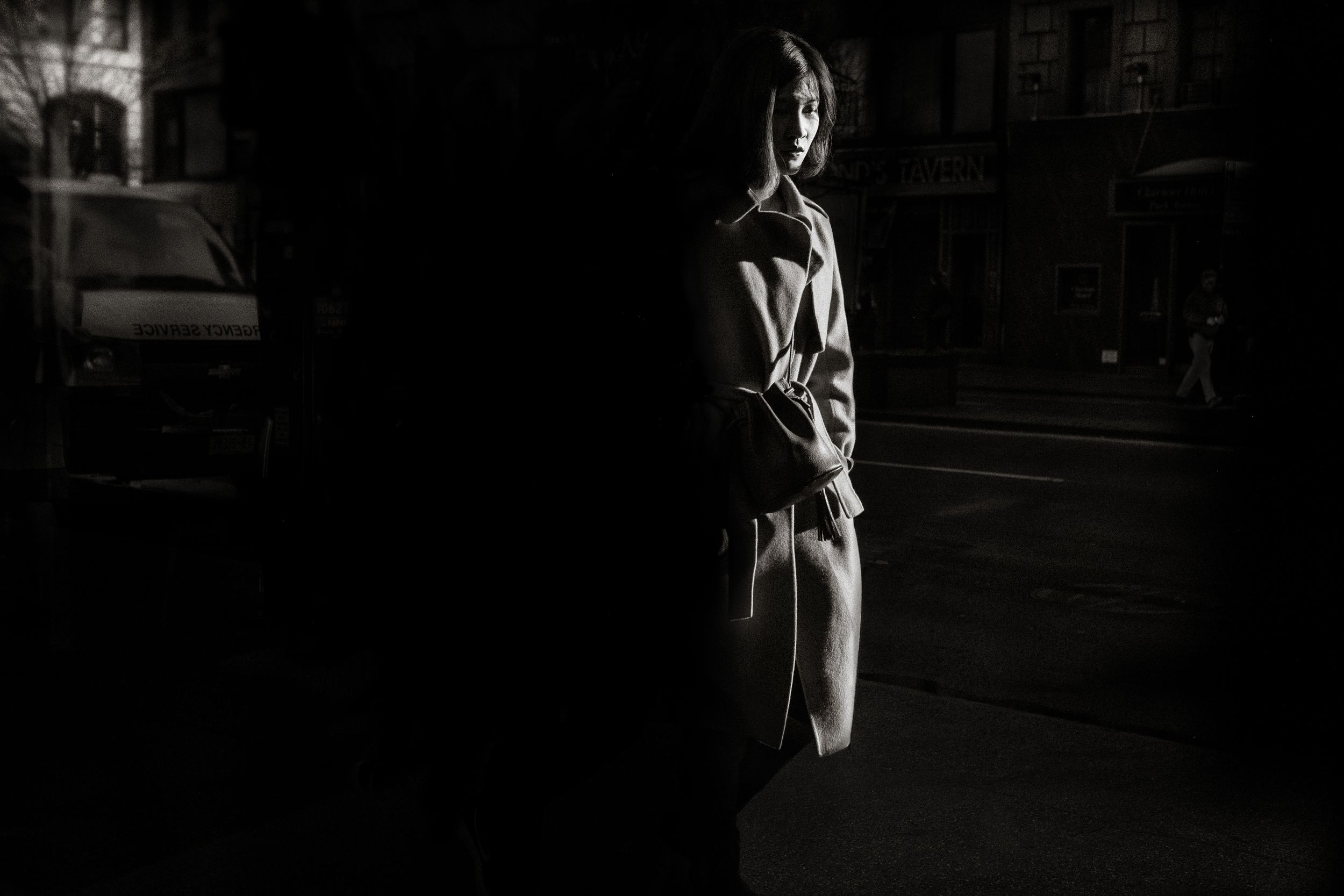 Black and white photo of a woman standing in urban shadows, leaning against a dark wall on a city street.