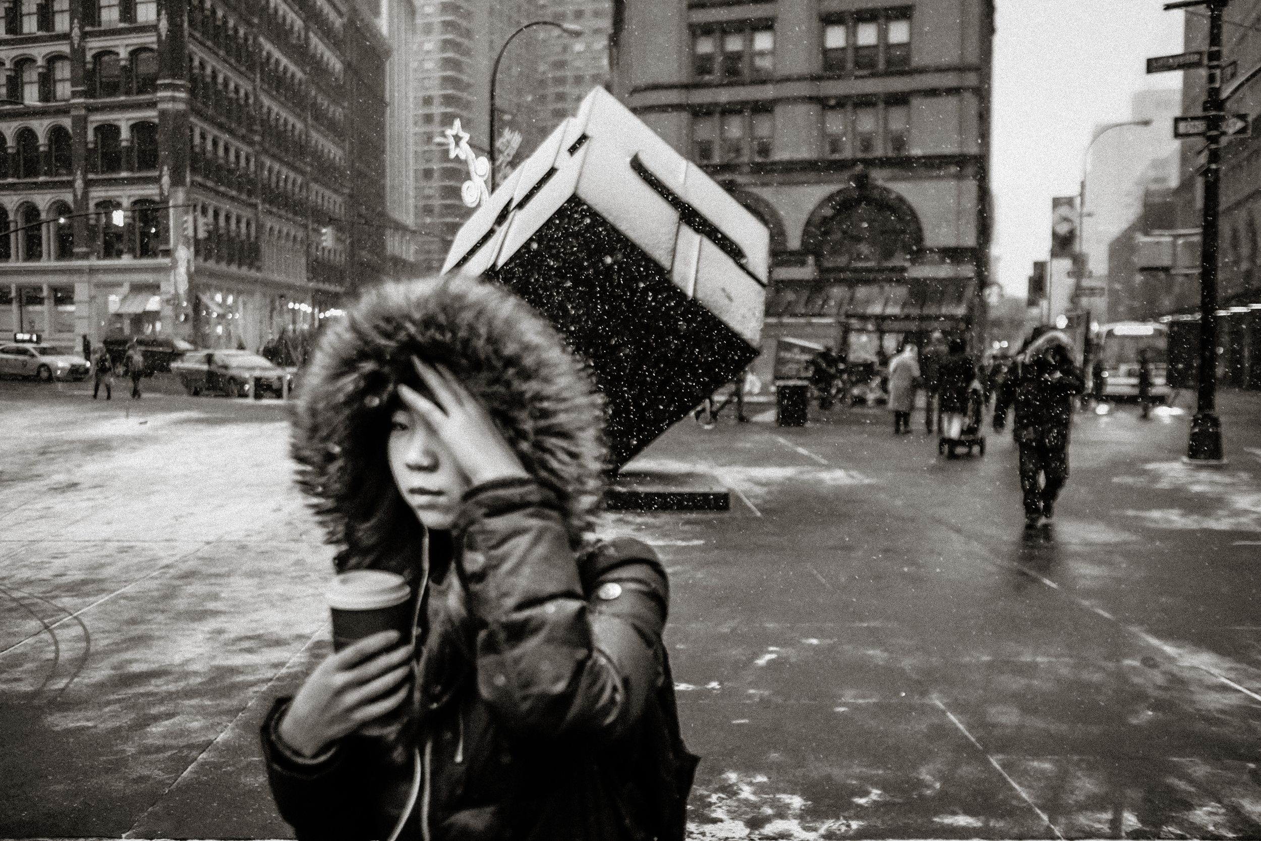 Black and white street photo of a woman in a fur-hooded coat carrying a box on her head in a snowy city.