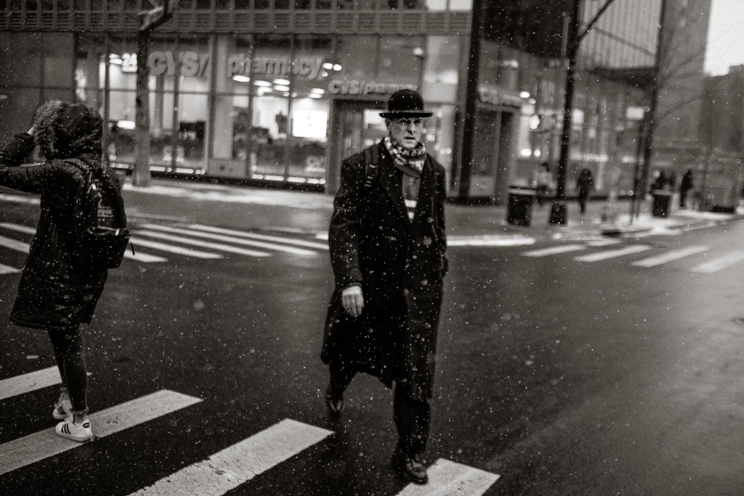Black and white street photo of an elderly man in a bowler hat crossing a snowy urban crosswalk at night.