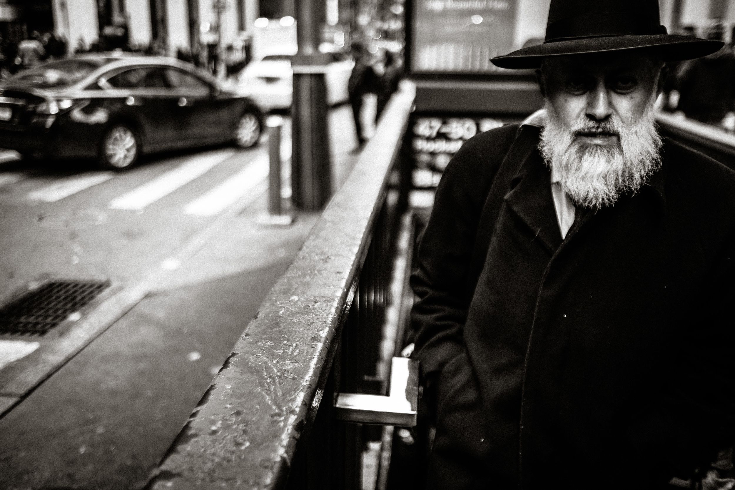 Black and white street photo of an elderly bearded man in a black hat and coat walking in an urban city setting.