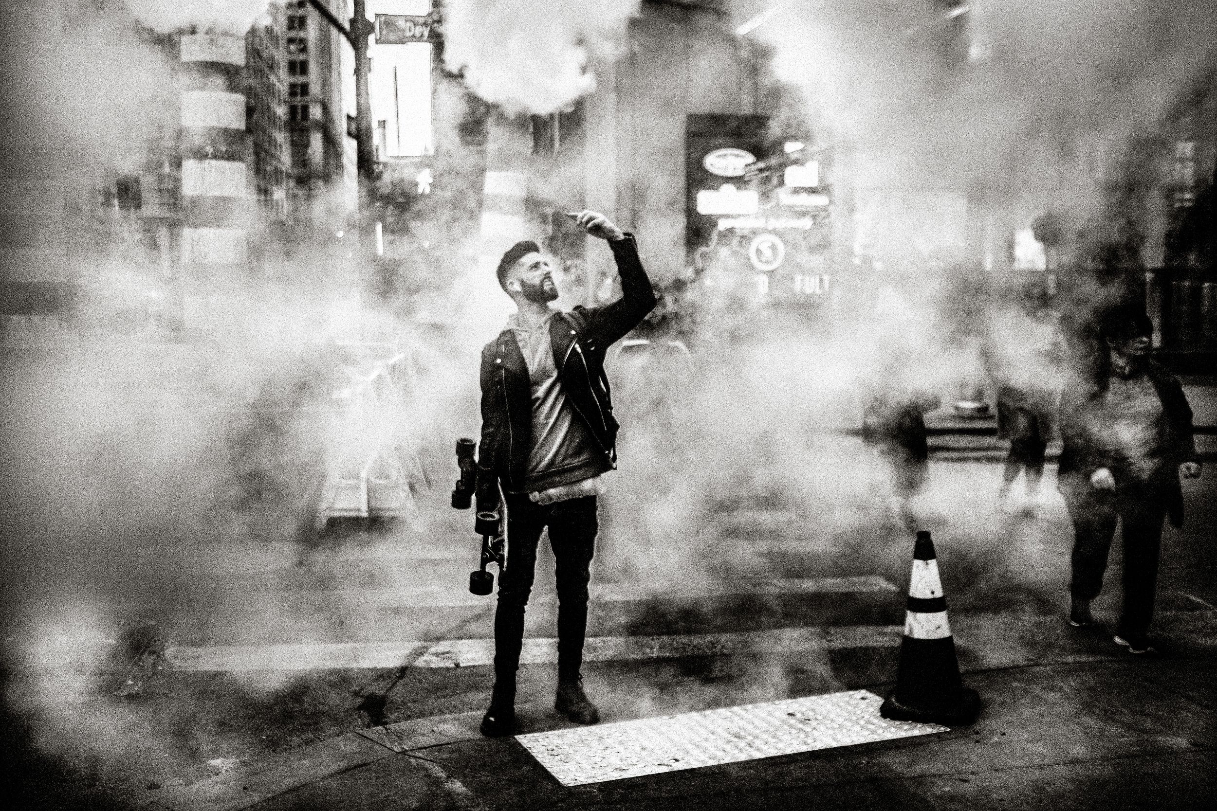 Black and white street photo of a man exhaling smoke surrounded by steam on a busy city street at night.