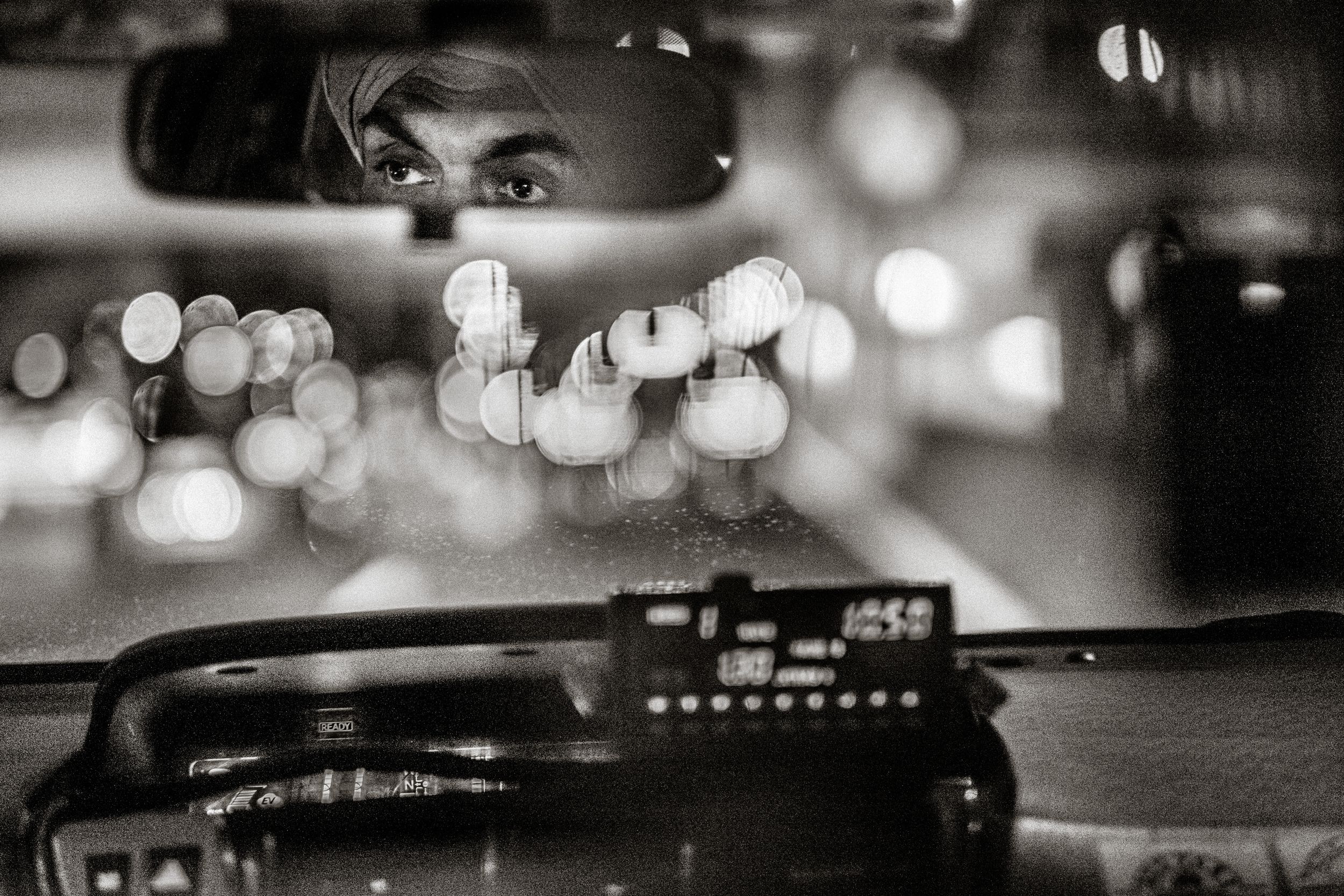 Black and white photo of a taxi driver reflected in rearview mirror with bokeh city lights at night.
