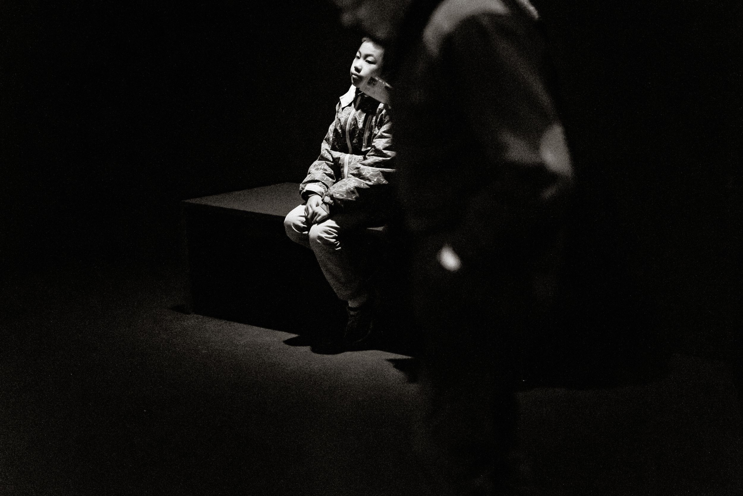 Black and white photo of a man sitting alone on a bench in dramatic spotlight against a dark background.