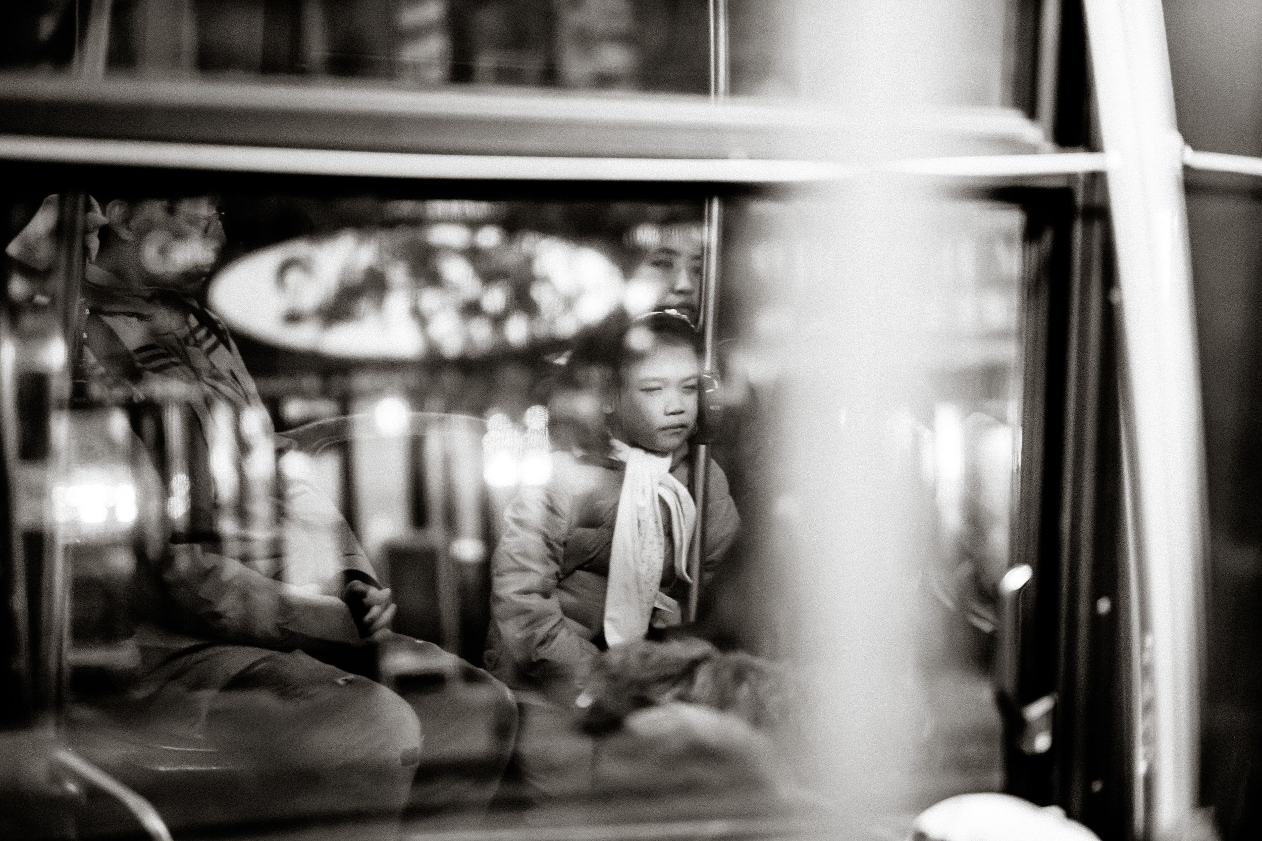 Black and white photo of a child sitting inside a bus, seen through the window with reflections.