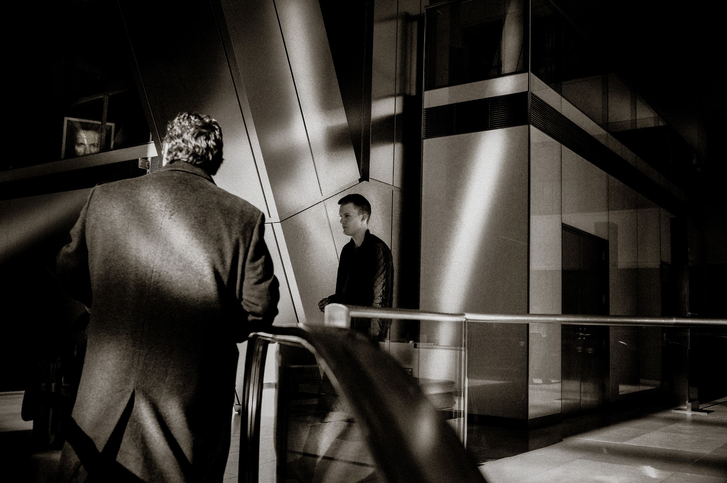Black and white photo of two men in a modern building with dramatic geometric shadows and glass walls.