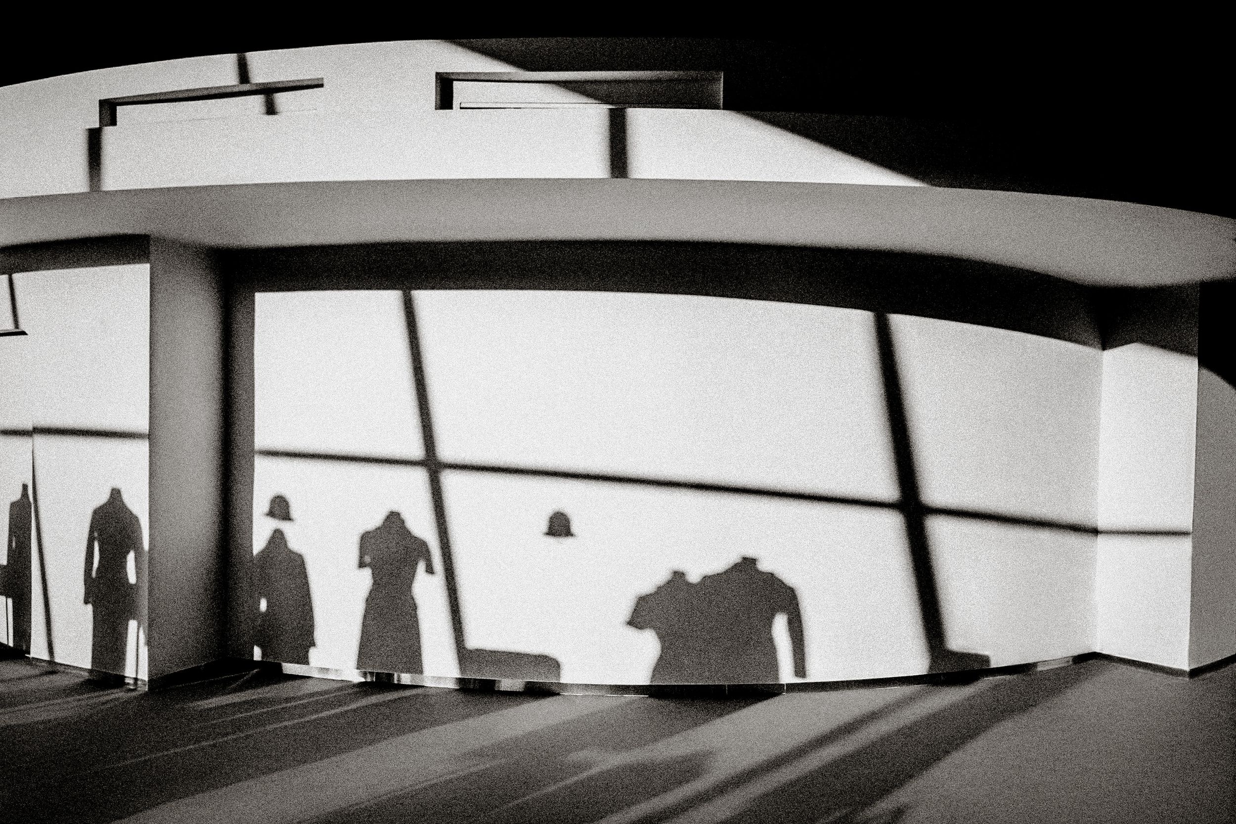 Black and white photo of dramatic shadows of three figures cast on a curved white building wall.