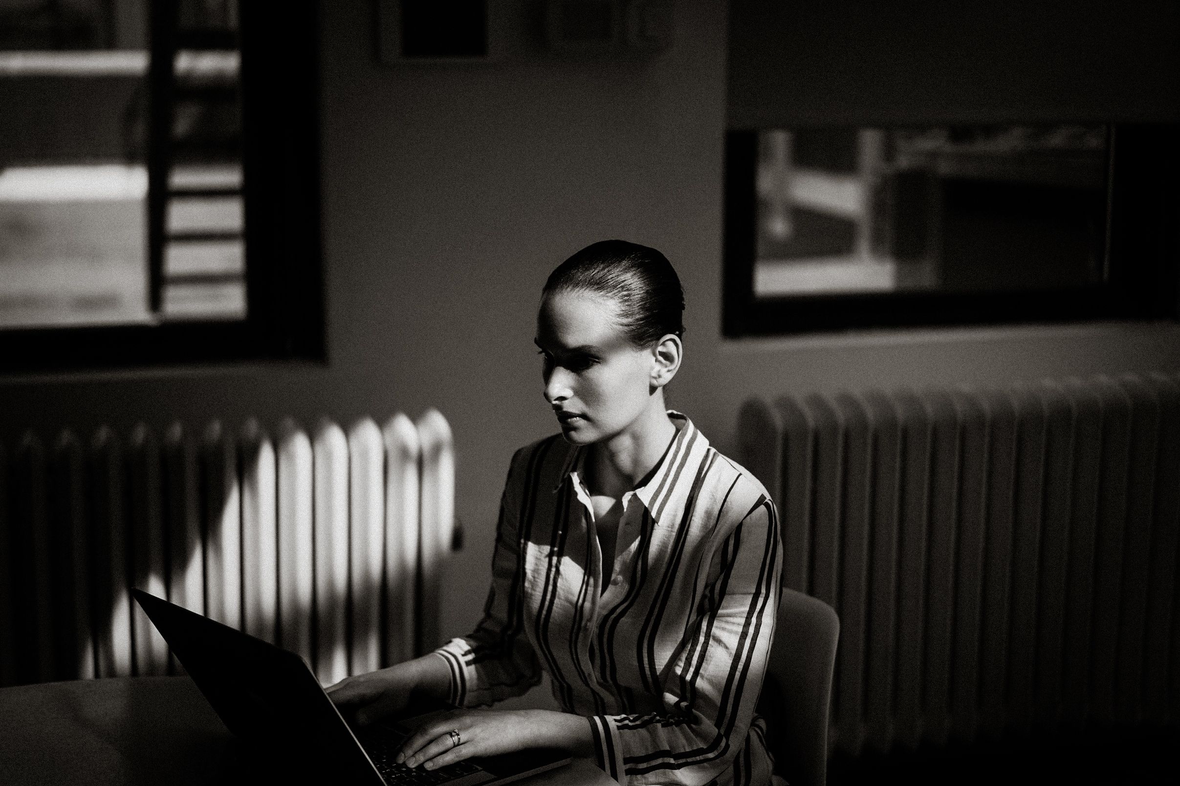 Black and white photo of a focused woman working on a laptop in a dimly lit room with a radiator behind her.