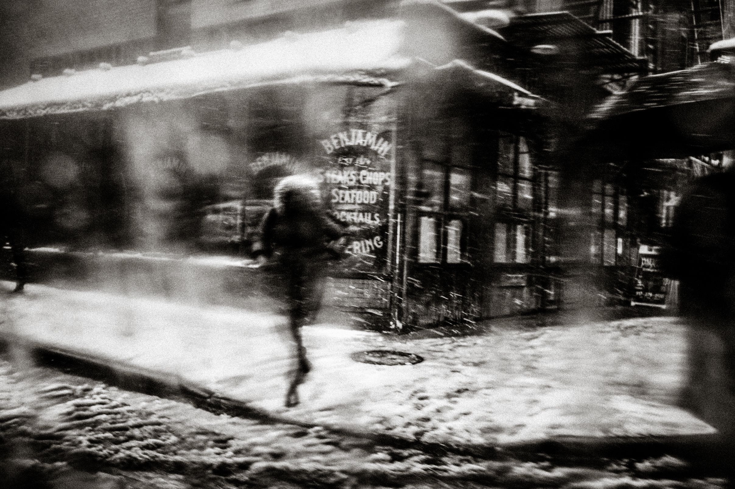 Blurry black and white photo of a person walking in a snowstorm past a lit restaurant at night.