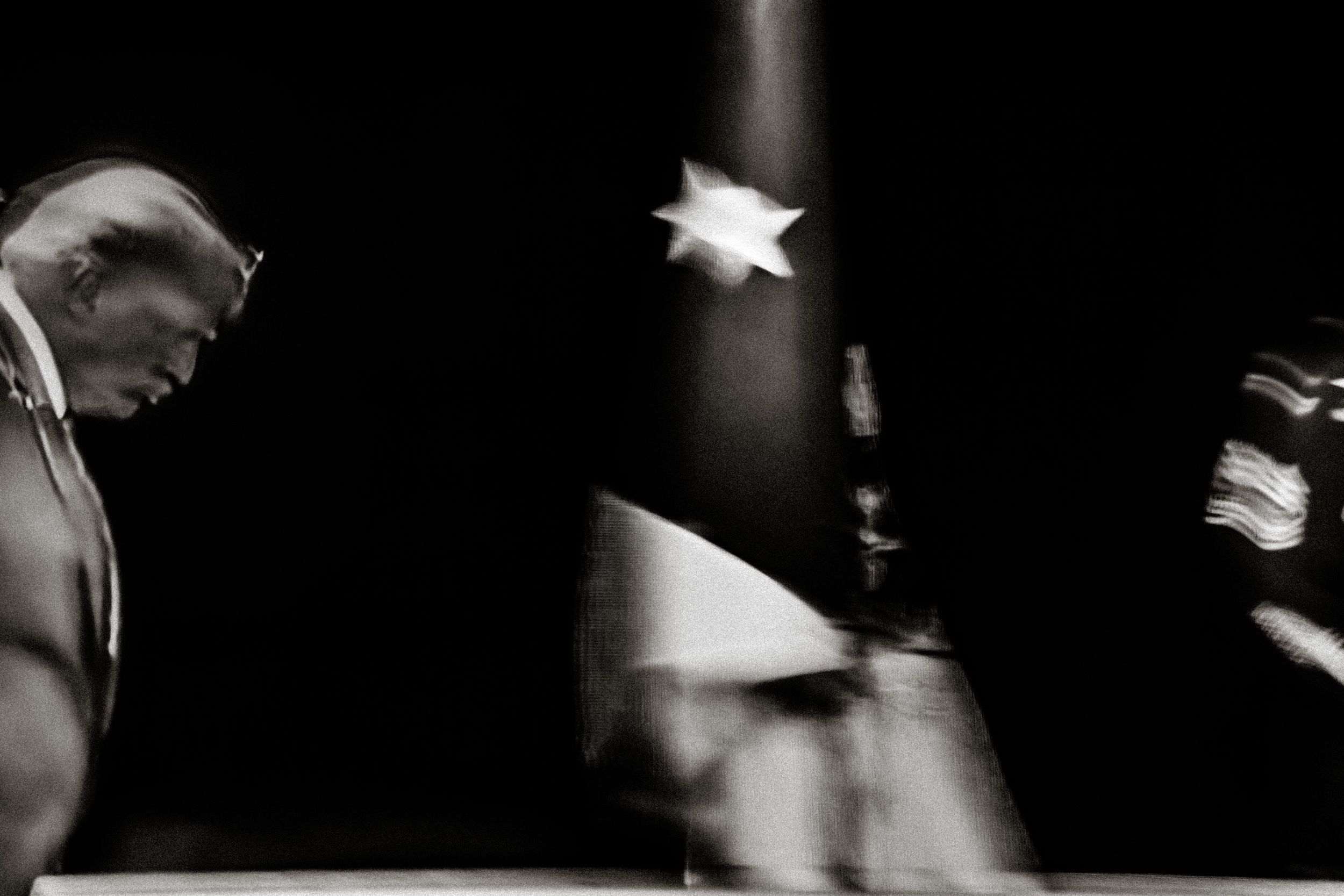 Black and white photo of a man in suit bowing head near a waving American flag in dramatic lighting.