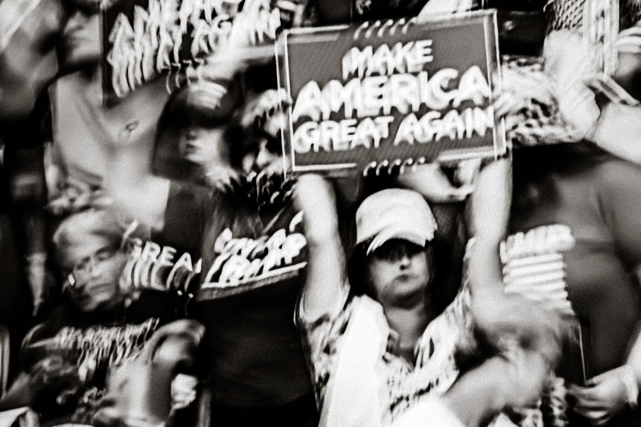 Black and white photo of crowd at political rally holding Make America Great Again signs.