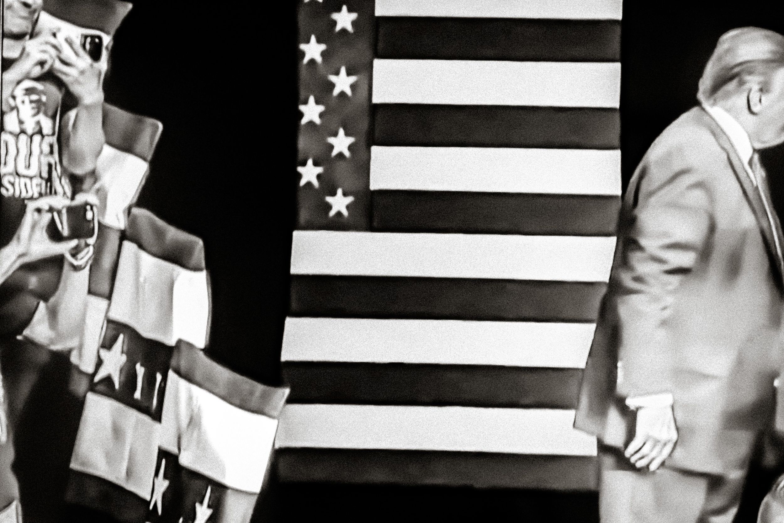 Black and white photo of two people standing before a large American flag at a political rally event.