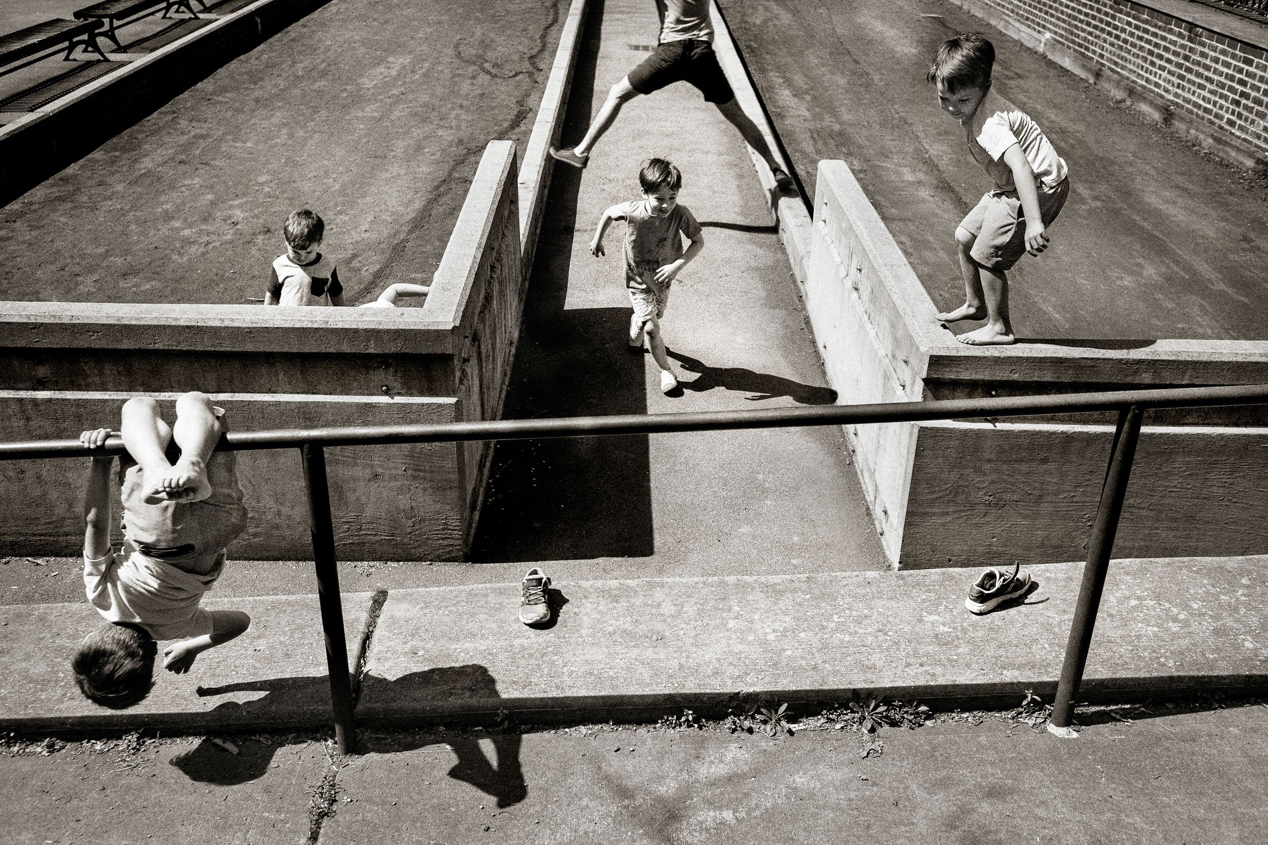 Black and white photo of children playing on a concrete skate park ramp, with shoes left on the ground nearby.