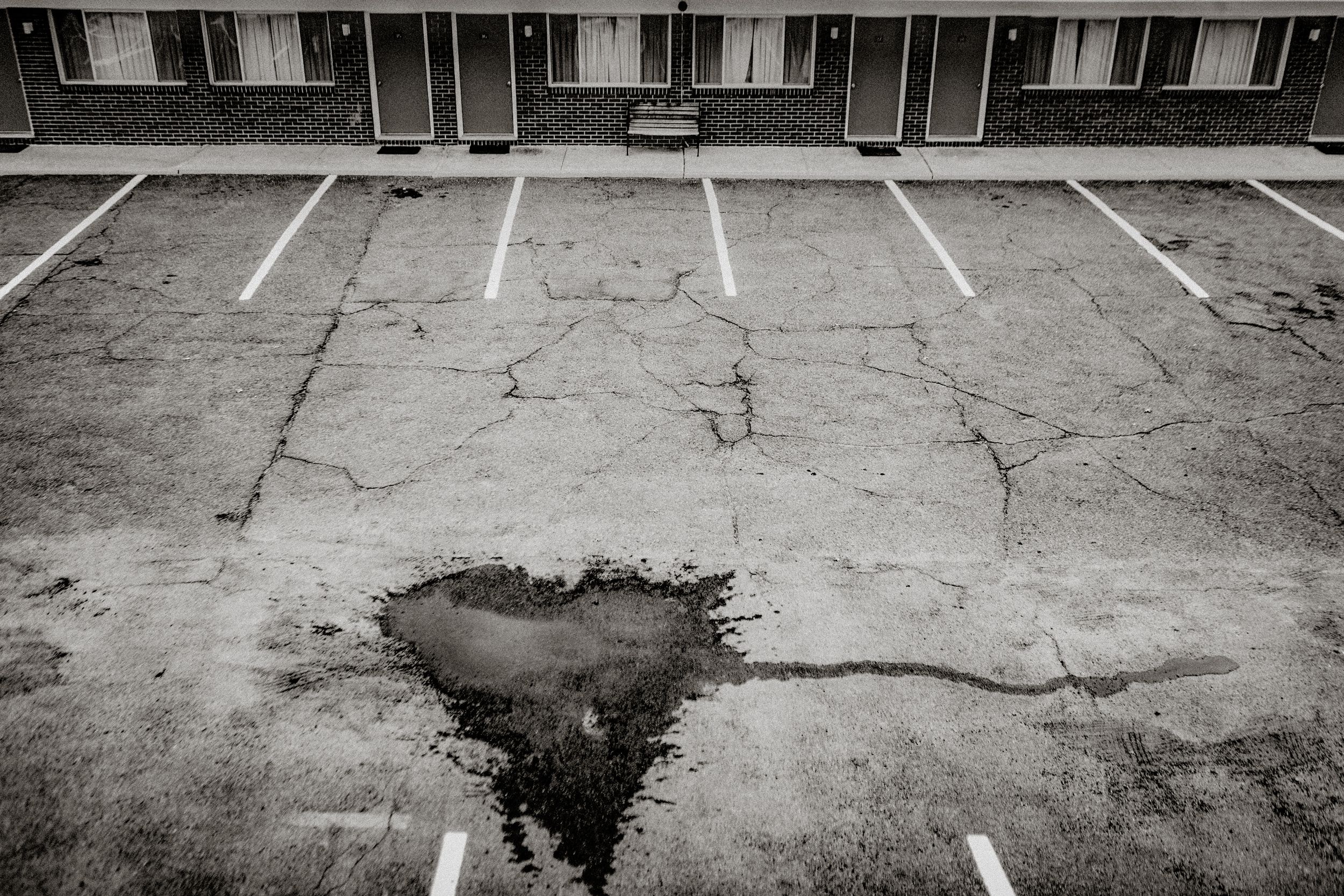 Black and white photo of cracked asphalt parking lot with puddle near motel building.