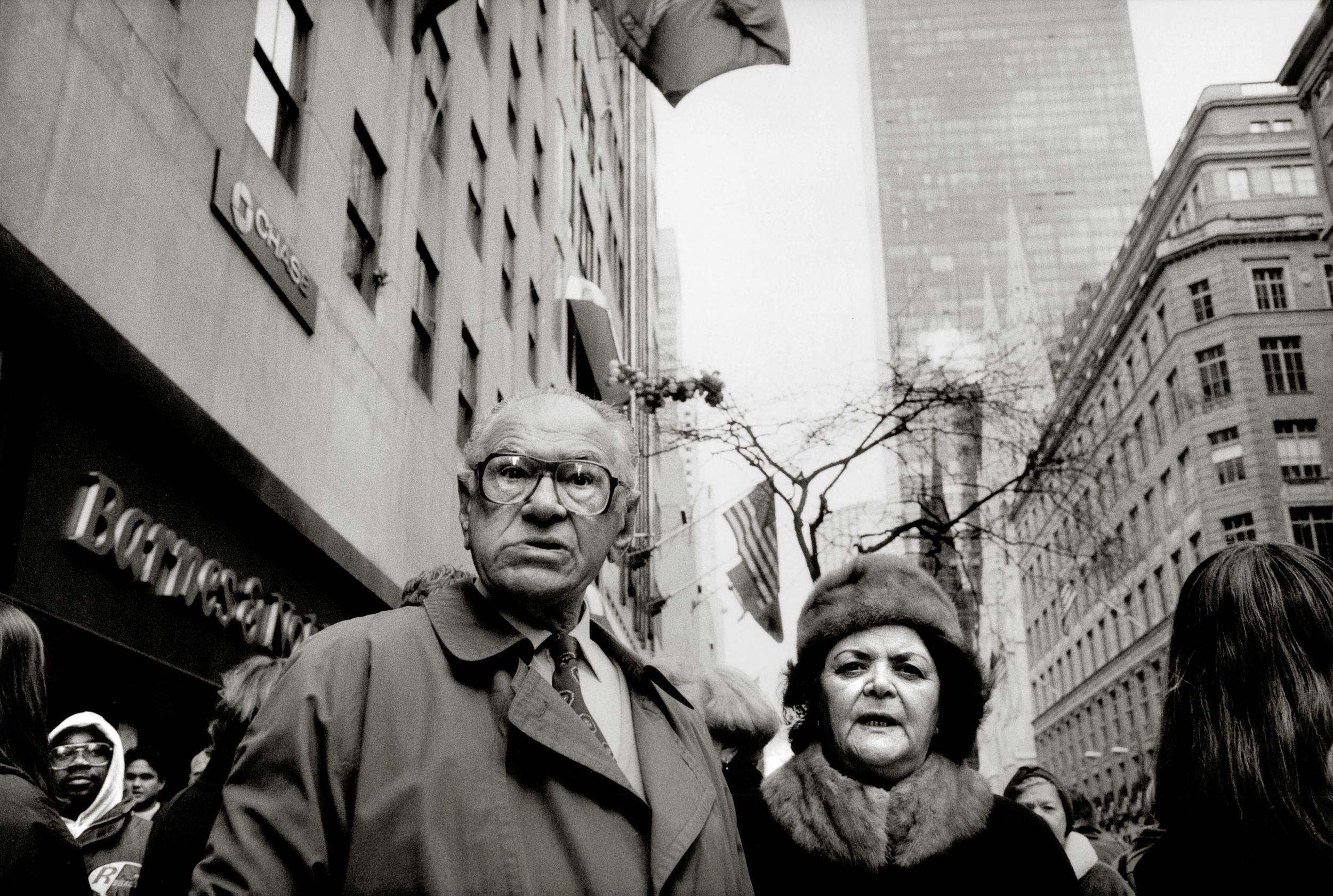 Black and white photo of an elderly couple standing on a busy city street with tall buildings in the background.