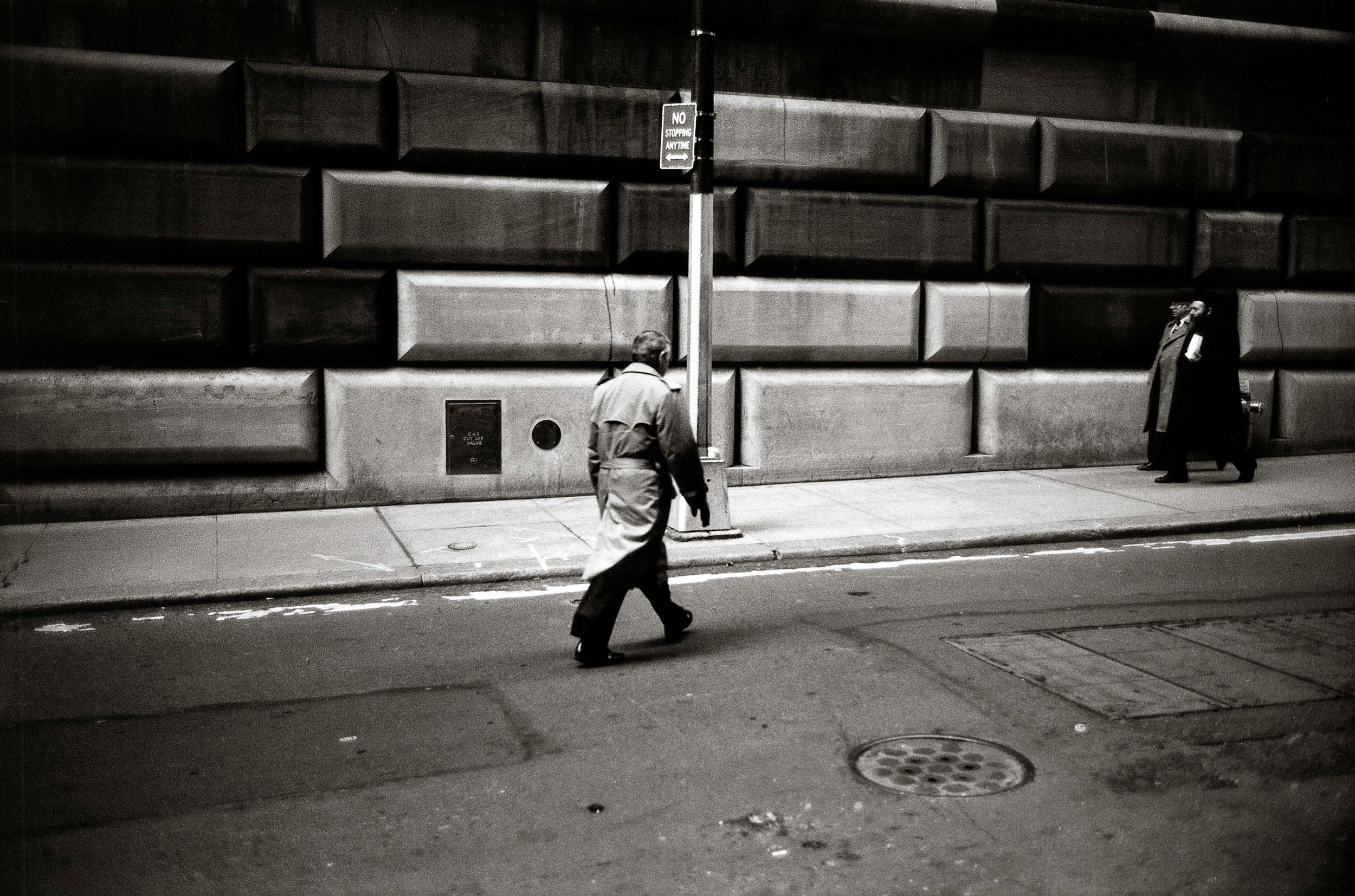 Black and white photo of a person walking on an urban sidewalk past a large stone building.
