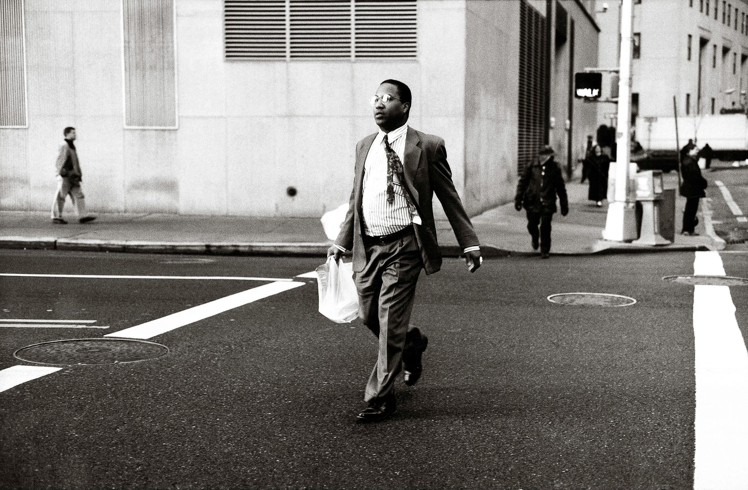 A Black man in a suit and tie crosses a city street carrying a white bag in a black and white urban street photo.