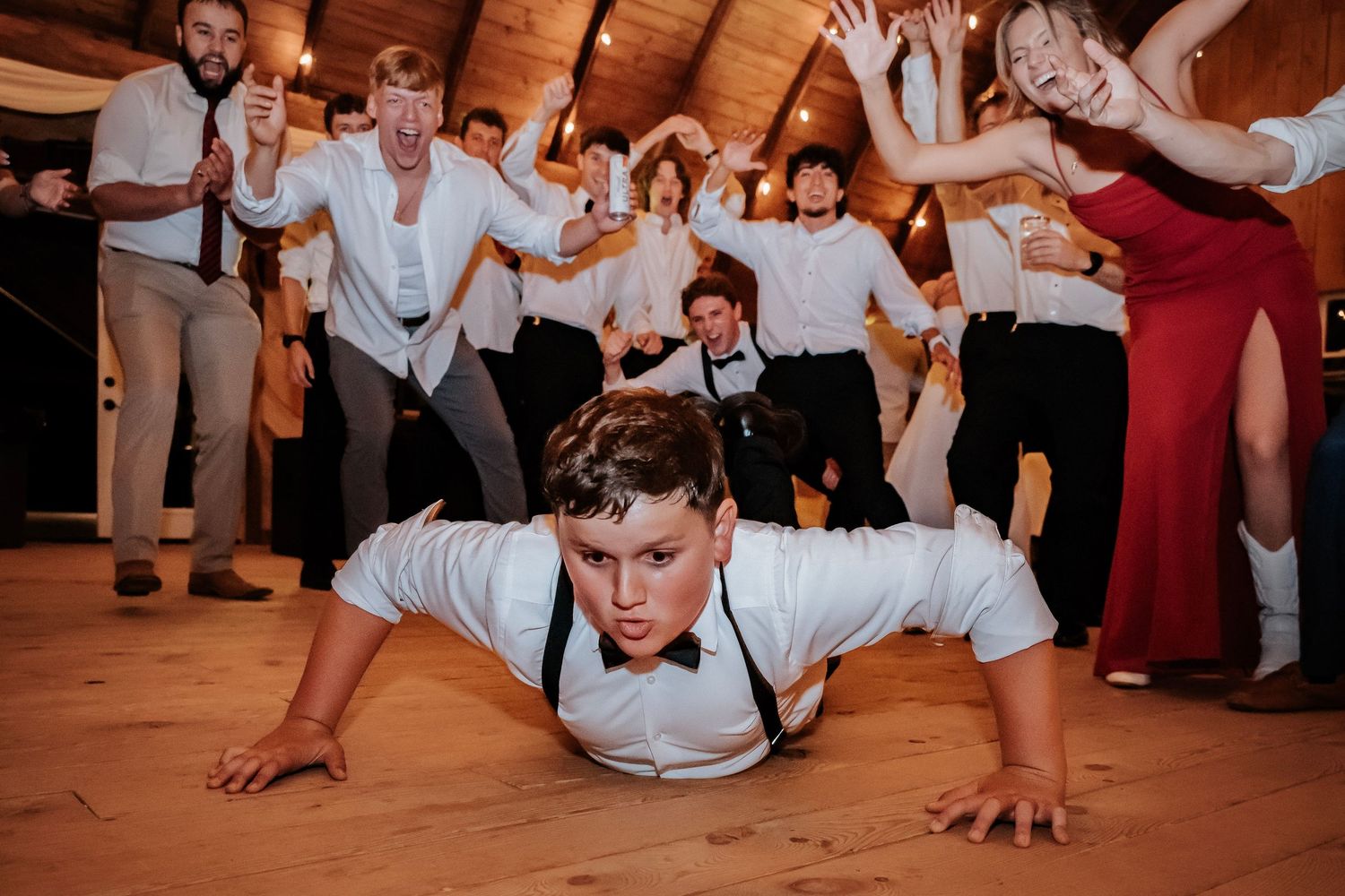 Boy doing push-ups on dance floor at wedding reception with cheering guests celebrating behind him.