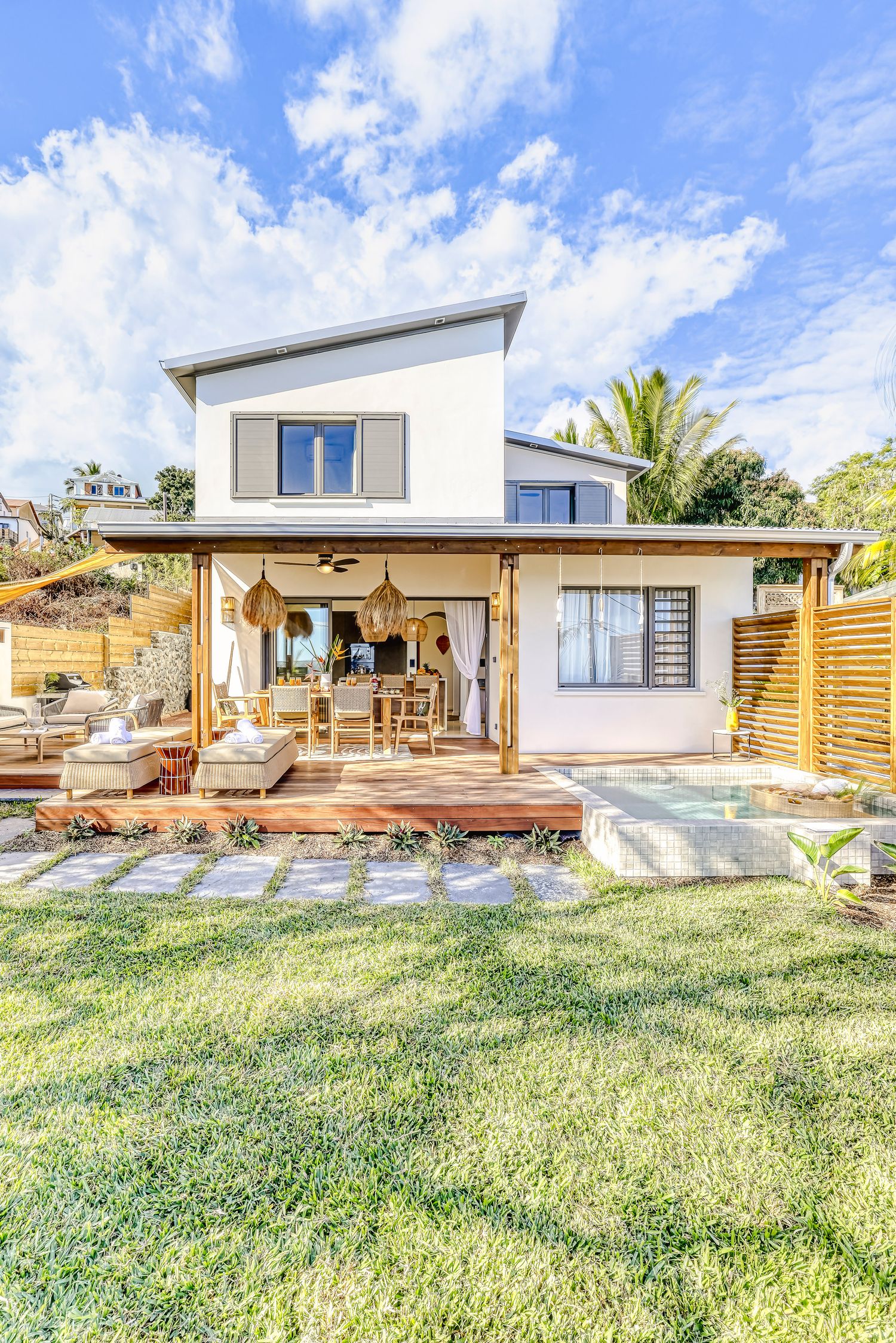 Modern two-story beach house with wooden deck, outdoor seating, and lush green lawn under blue sky.