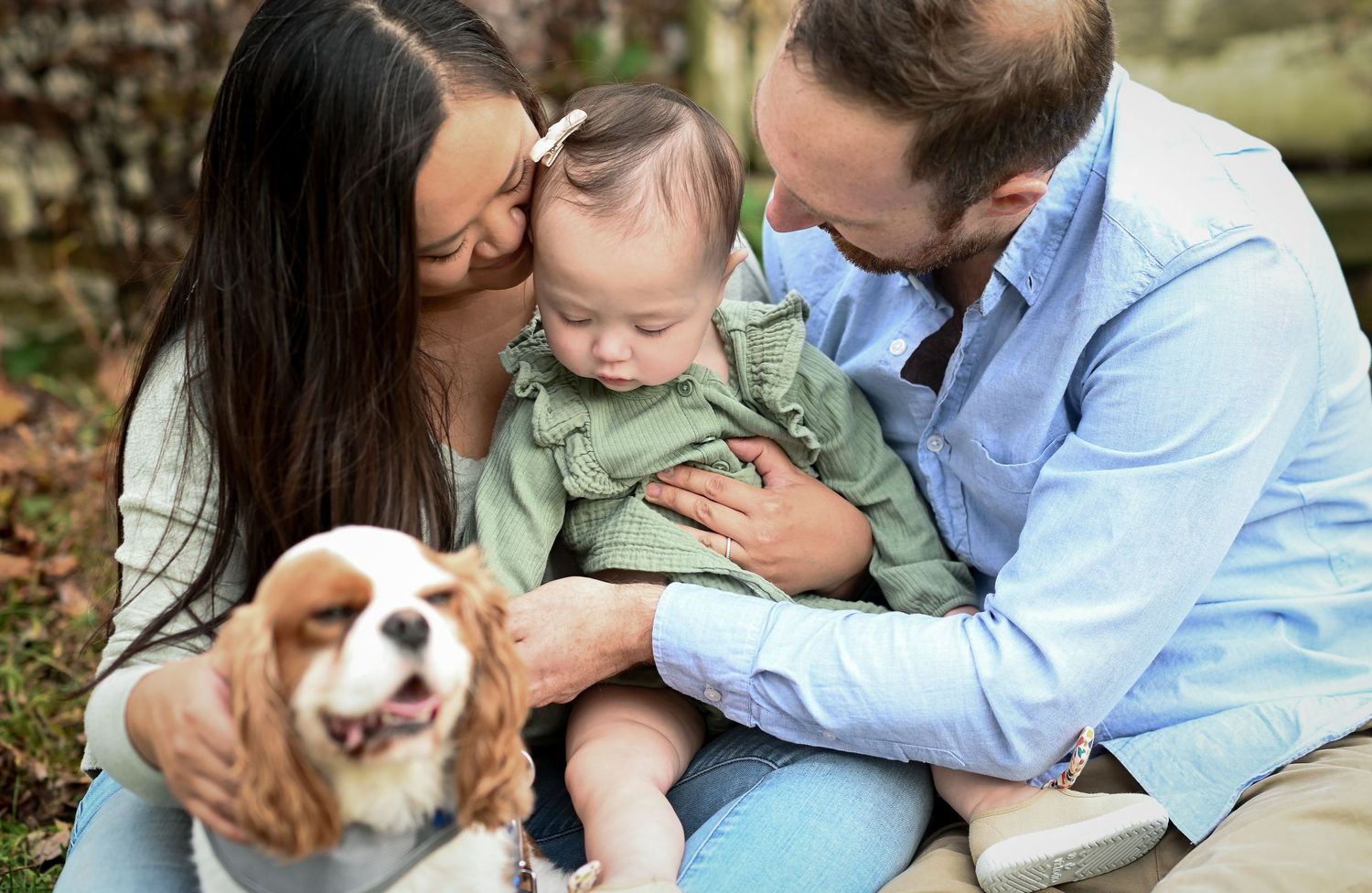A young family with a baby girl and a Cavalier King Charles Spaniel posing outdoors in a warm, candid portrait.