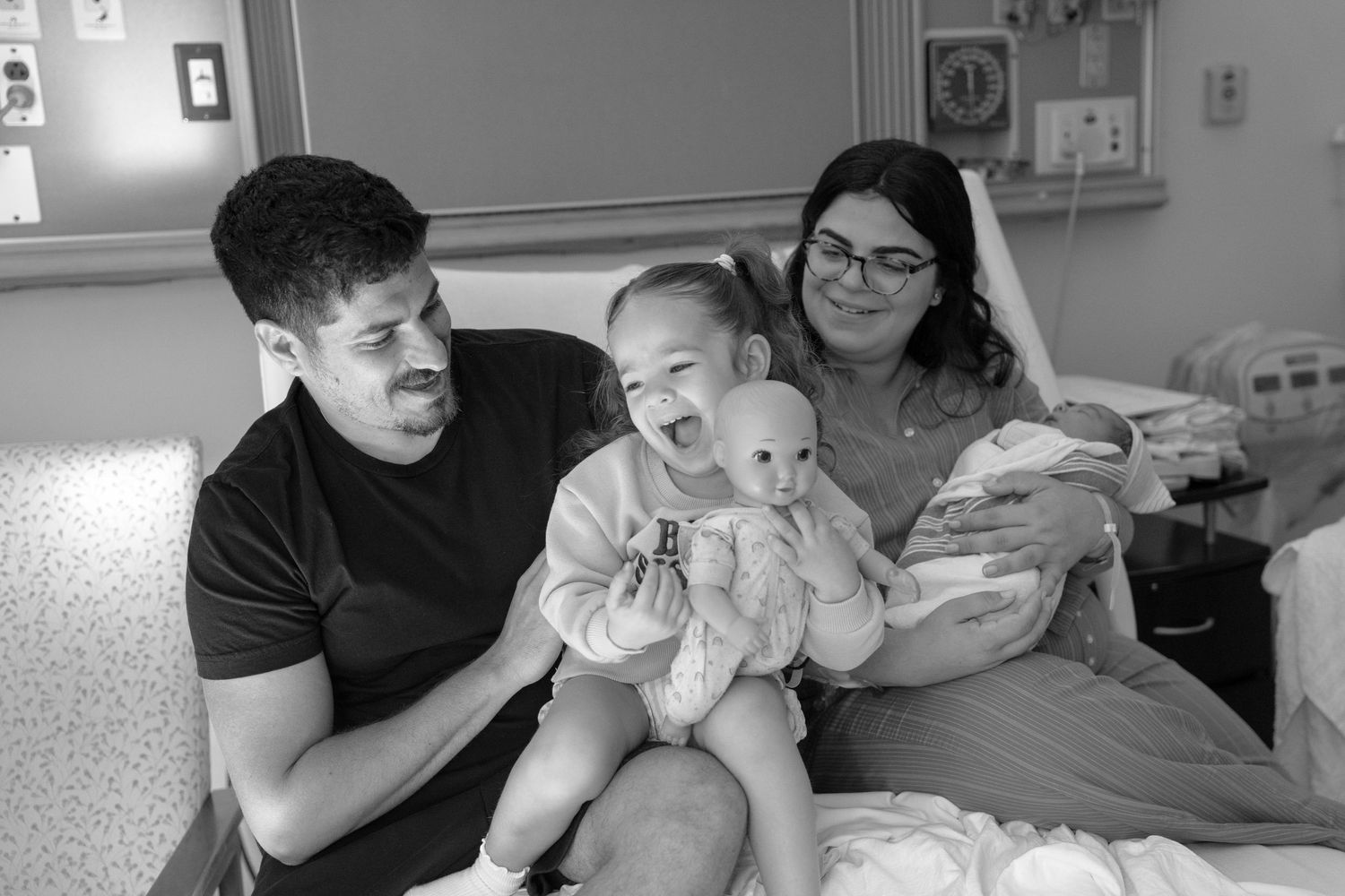 Black and white hospital photo of parents with toddler and newborn baby, family smiling together after birth.