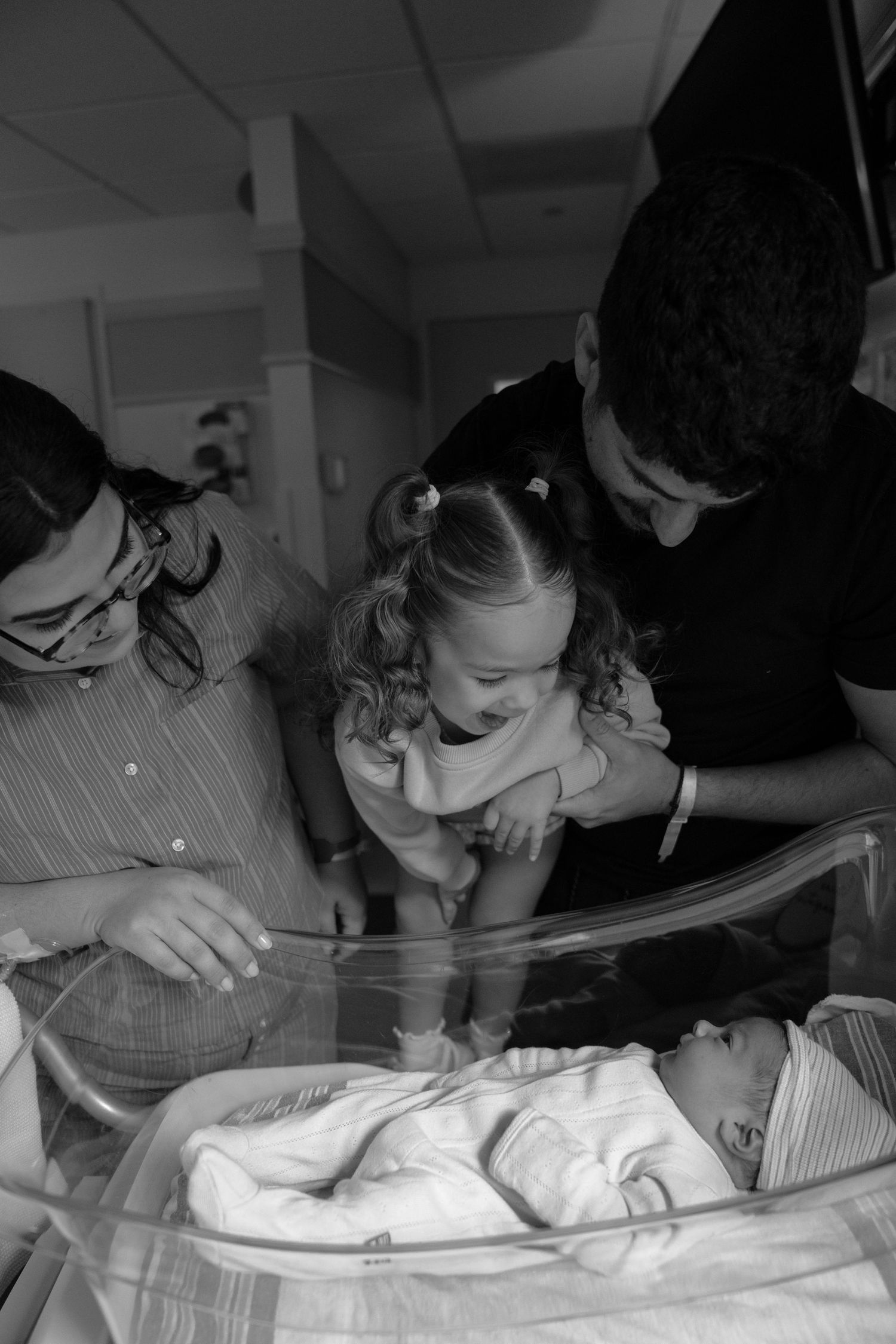 Black and white photo of parents and toddler girl meeting newborn baby in hospital bassinet.