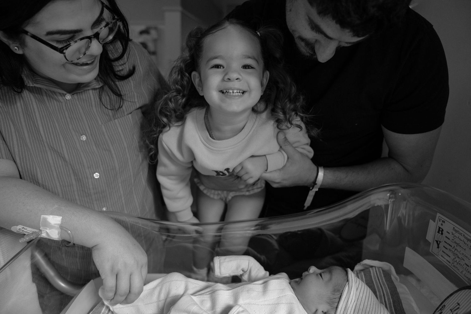 Black and white photo of a smiling toddler girl meeting her newborn sibling in hospital bassinet with parents nearby.