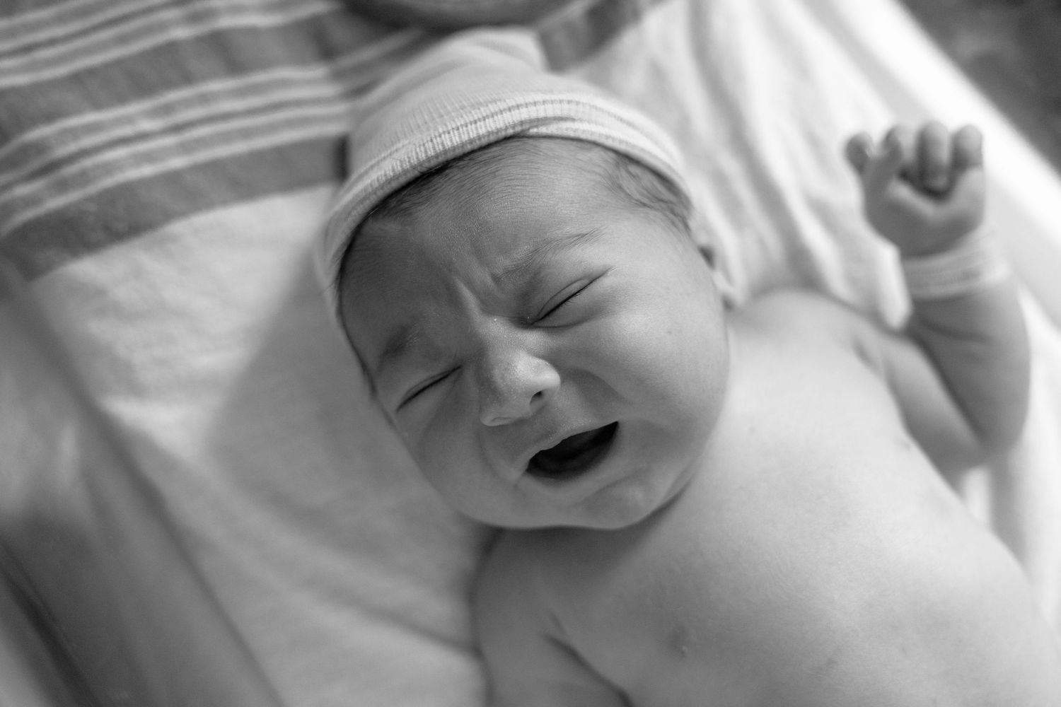 Black and white photo of a crying newborn baby wearing a knit hat, lying on a soft blanket.