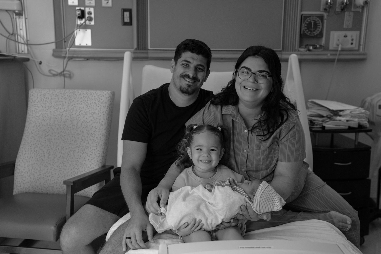 Black and white photo of a family with two parents and a toddler posing in a hospital room with a newborn baby.