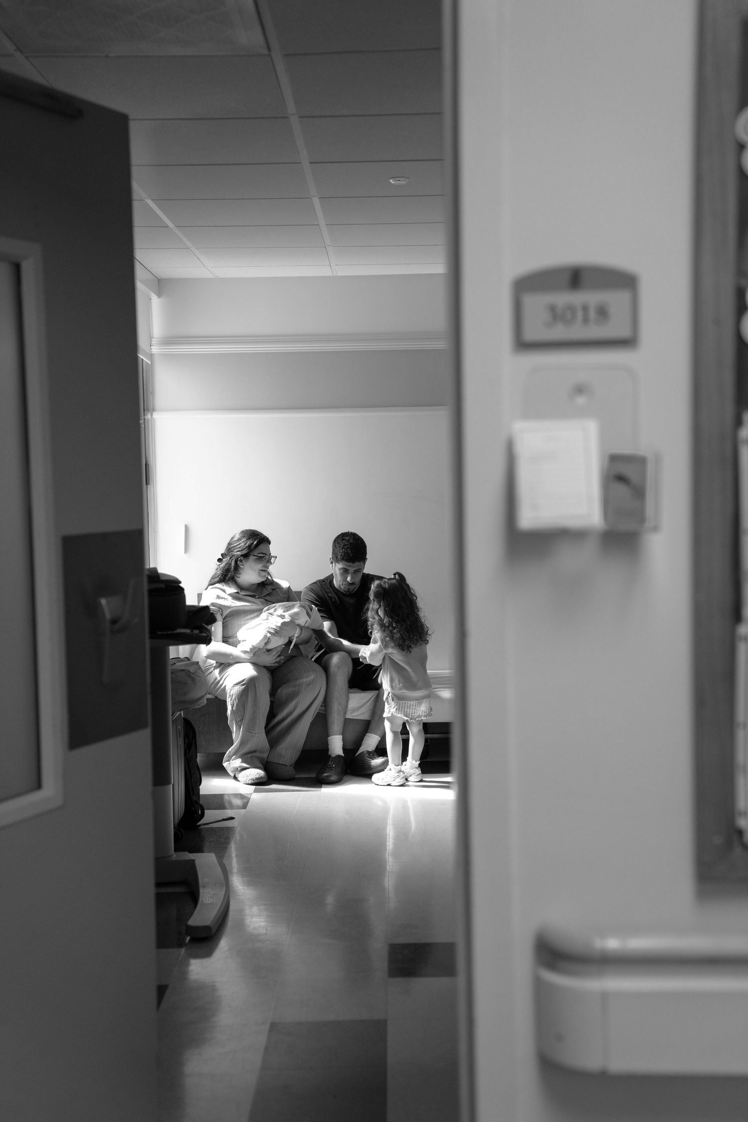 Black and white photo of a family sharing a tender moment in hospital room 3018, viewed through a doorway.