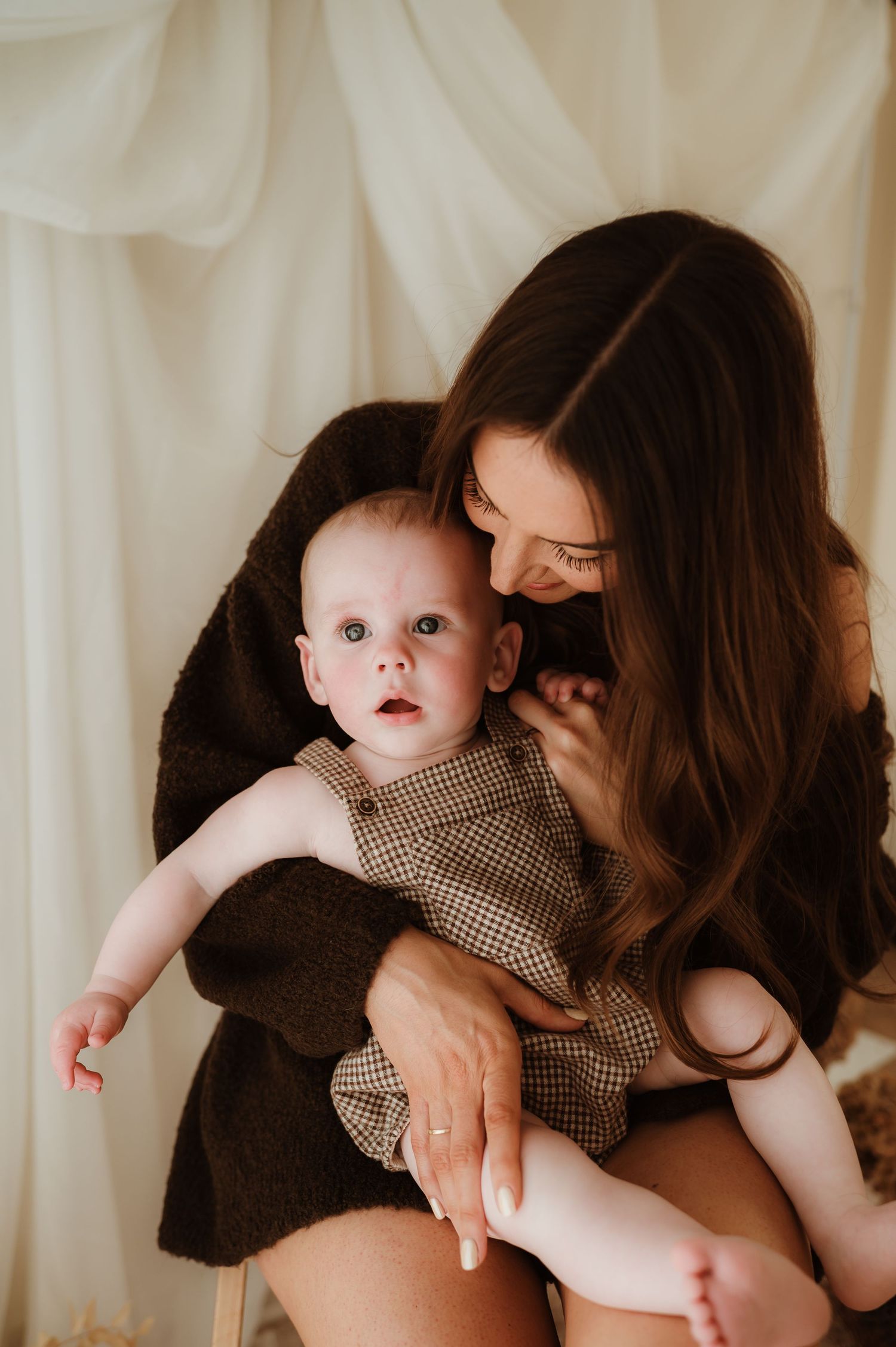A baby wearing a white ruffled dress sits on a textured cream blanket in warm natural lighting.