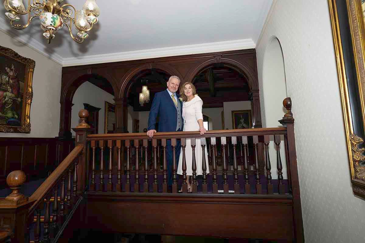 Elegant couple standing on a grand wooden staircase landing in a classic Victorian-style home interior.