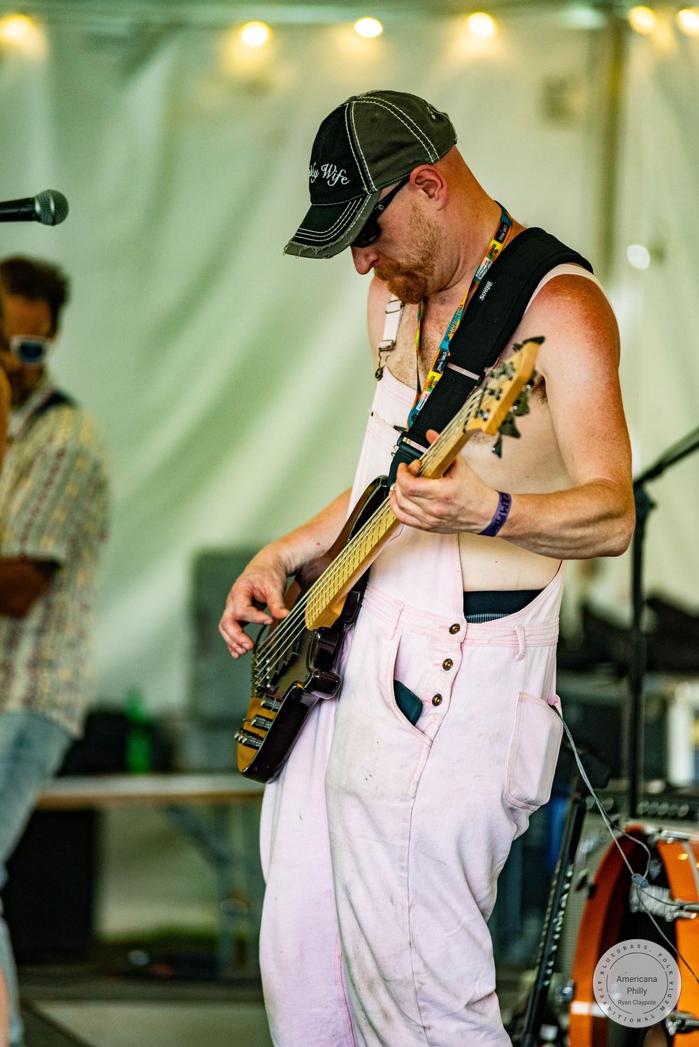 Shirtless male guitarist in white overalls and baseball cap performing on stage at an outdoor music festival.