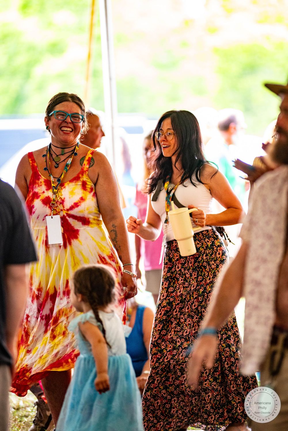 Two women laughing and dancing at an outdoor festival, one in a tie-dye dress, the other in a floral skirt.
