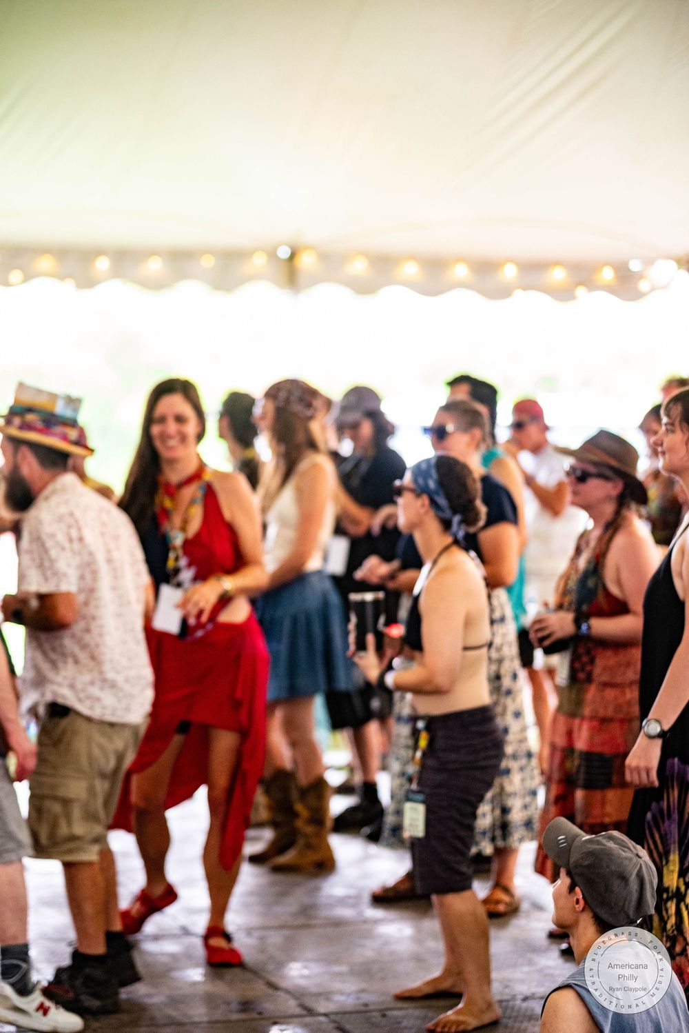 Crowd of festival-goers dancing under a tent with string lights, wearing colorful summer outfits and hats.