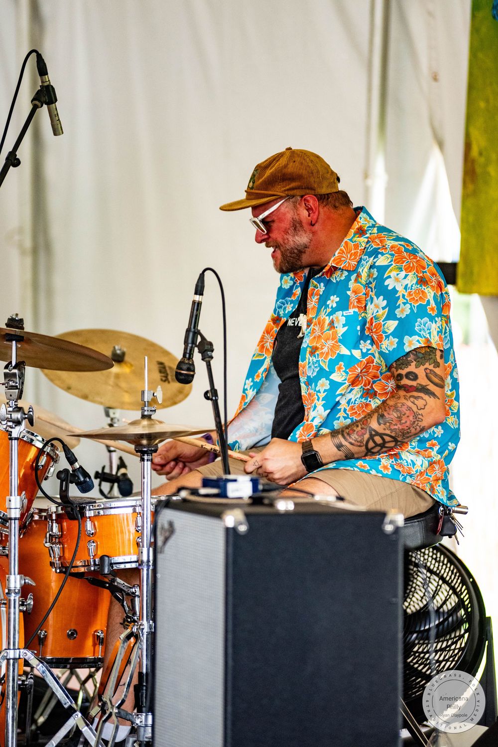 Tattooed drummer in floral shirt and cap playing drums on outdoor stage at music festival.