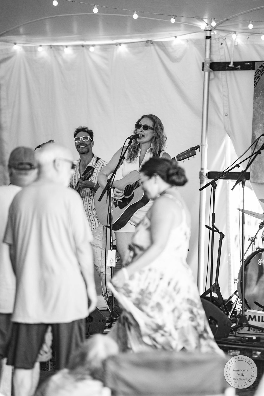 Black and white photo of a woman singing and playing acoustic guitar on stage under a tent with string lights at an outdoor event.