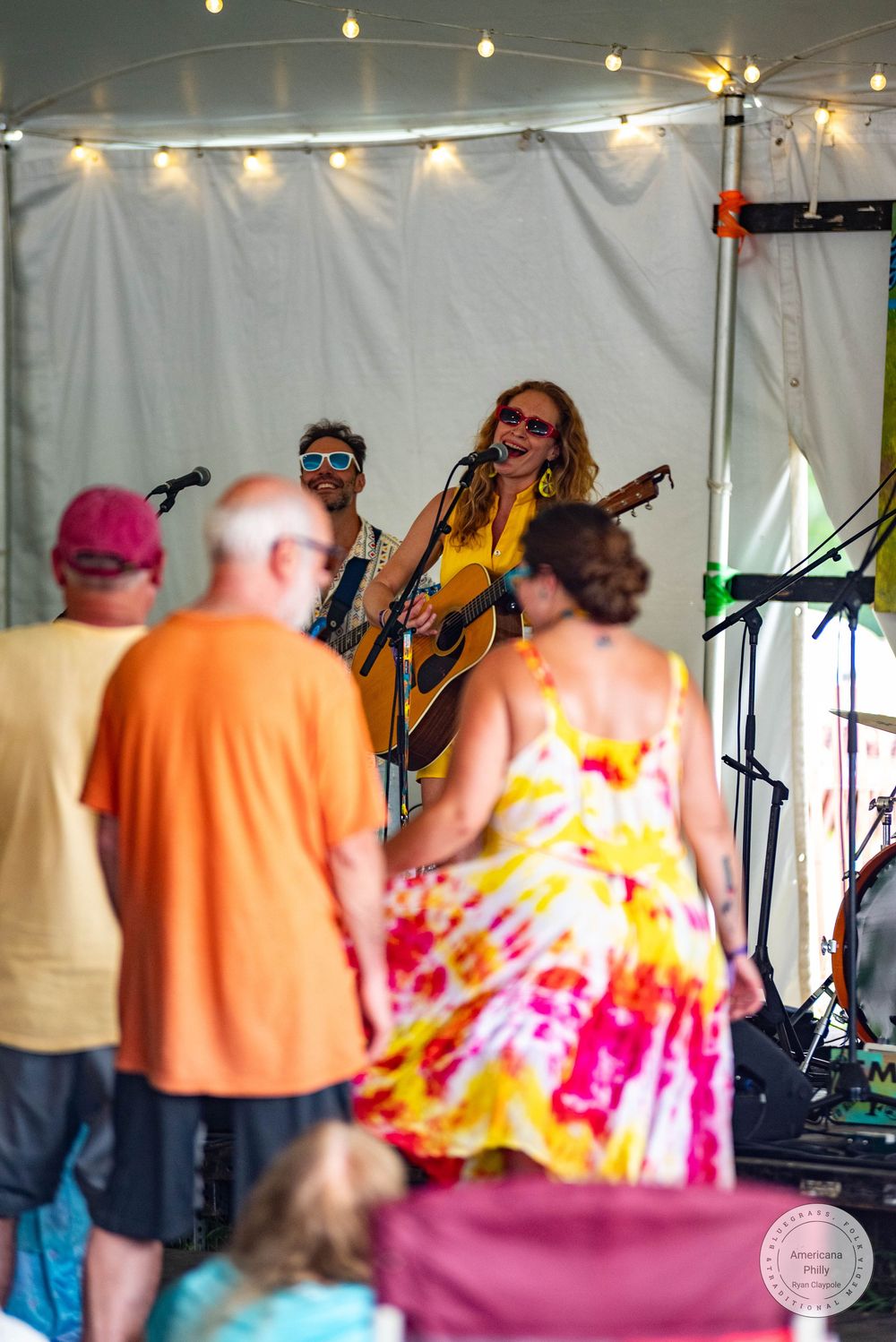 Live outdoor music festival performance with two singers playing acoustic guitars under a tent with string lights.