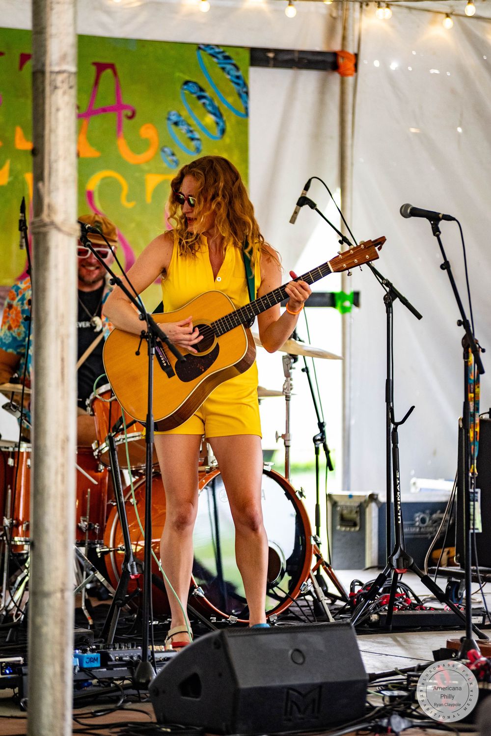 Woman in yellow romper playing acoustic guitar on stage at outdoor music festival under white tent.