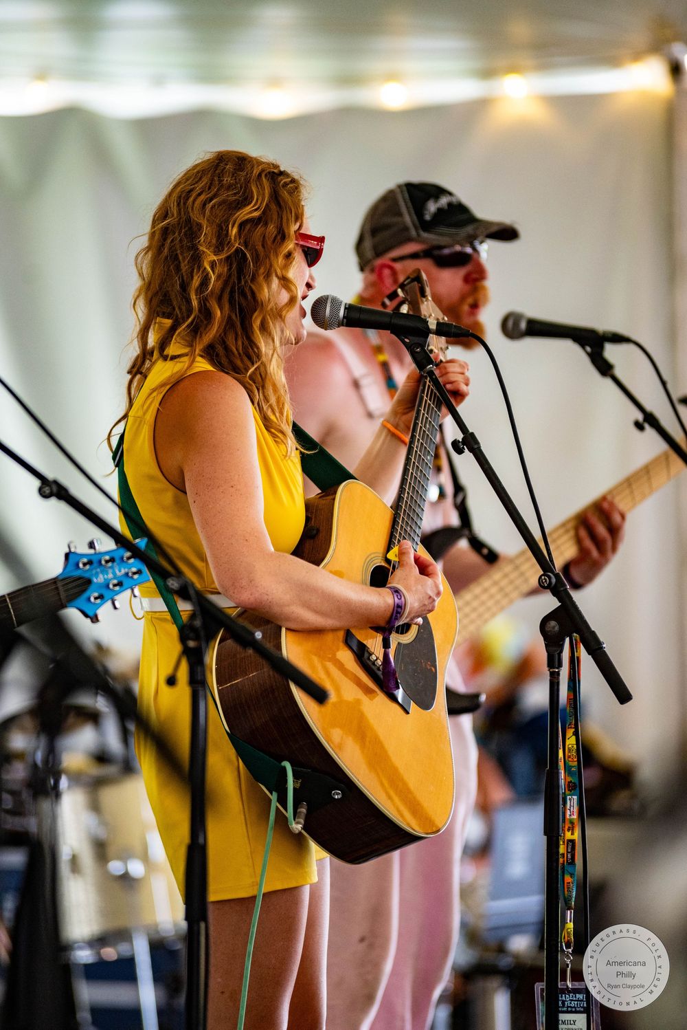 Woman in yellow dress plays acoustic guitar and sings at outdoor music festival while band performs behind her.