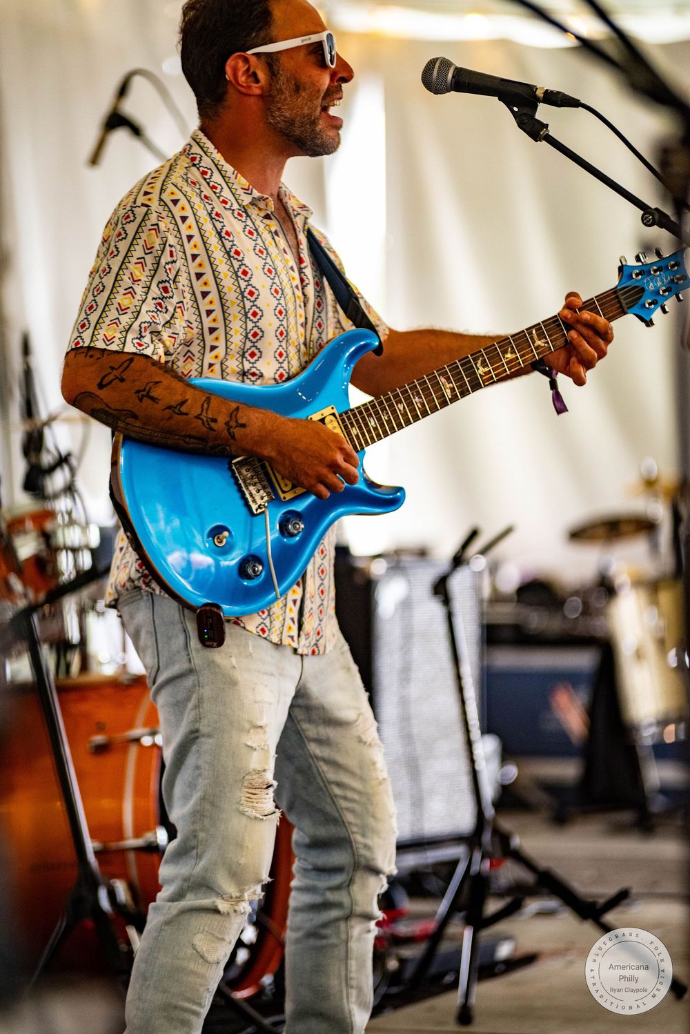Tattooed male guitarist playing blue electric guitar on stage, wearing patterned shirt and ripped jeans, performing at outdoor music event.