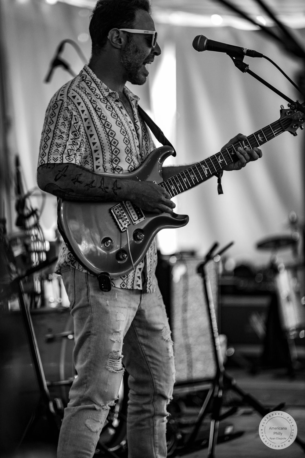 Black and white photo of a tattooed male guitarist performing on stage with a PRS guitar and microphone stand.